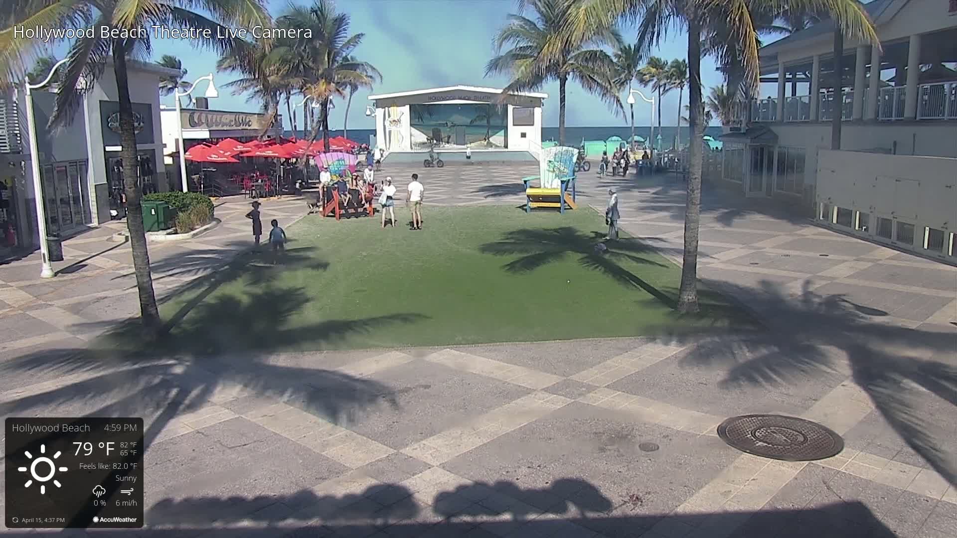 A sunny outdoor scene shows a paved plaza with a grassy area, palm trees, a beachfront stage, and several people walking around.