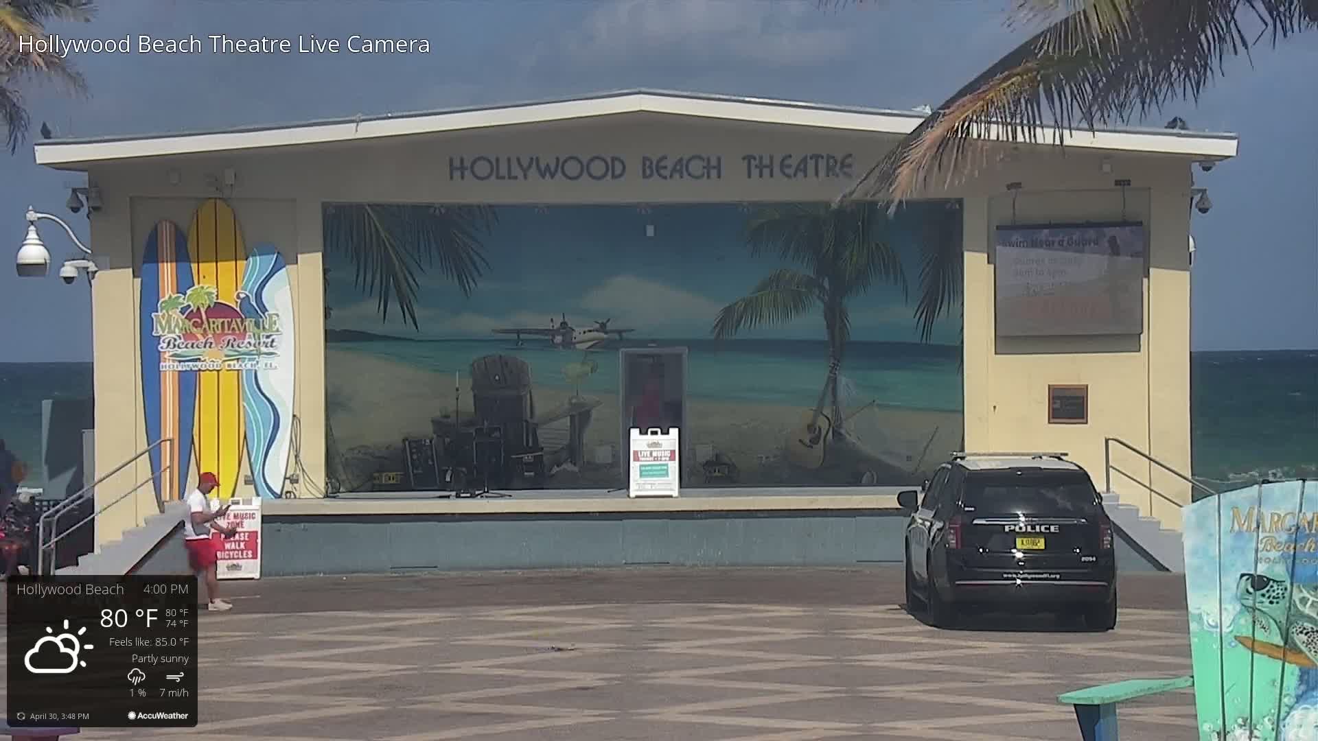 A partly sunny beach scene shows an outdoor stage with a tropical mural, a police car parked in front, and two surfboards leaning against the stage.