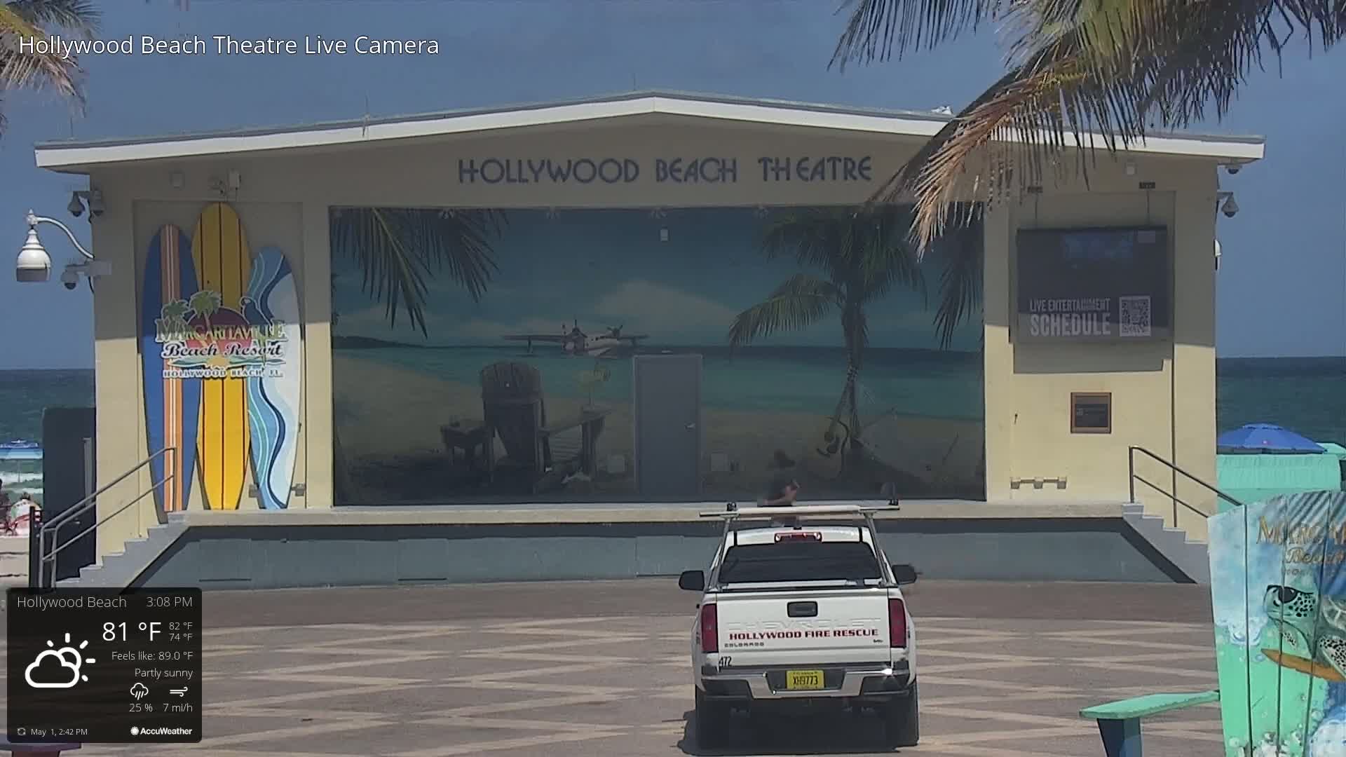 A partly sunny beachside stage with a mural, a fire rescue truck parked in front, and a calm ocean in the background.