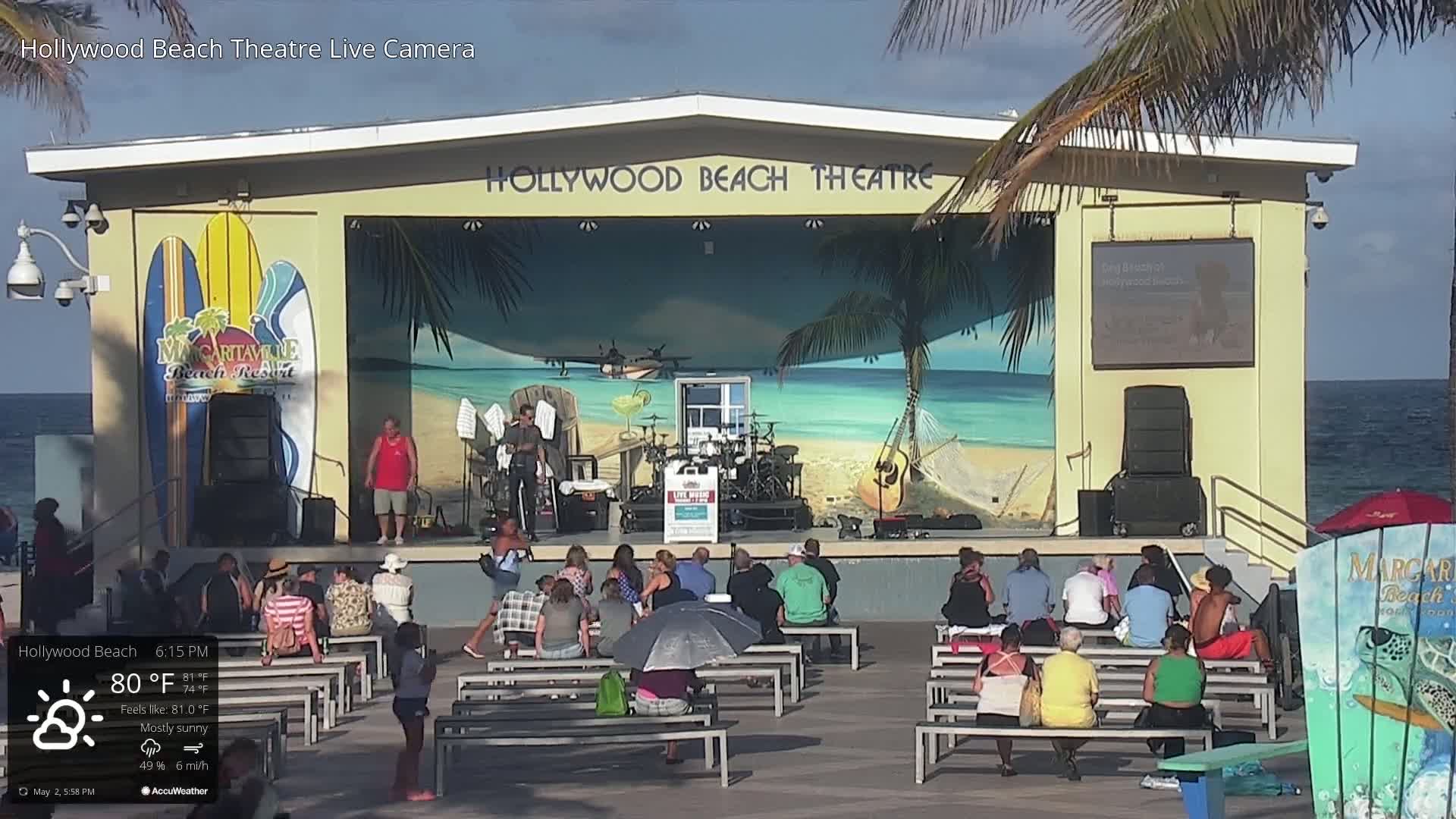 An outdoor stage with a band playing, audience members seated in front, and a mostly sunny sky.