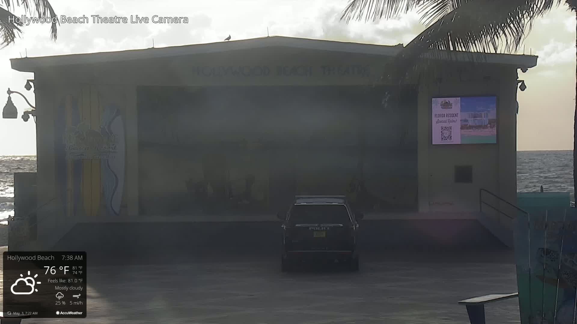 A mostly cloudy morning reveals a dark-colored police vehicle parked in front of a beachside theater, with the ocean visible in the background.