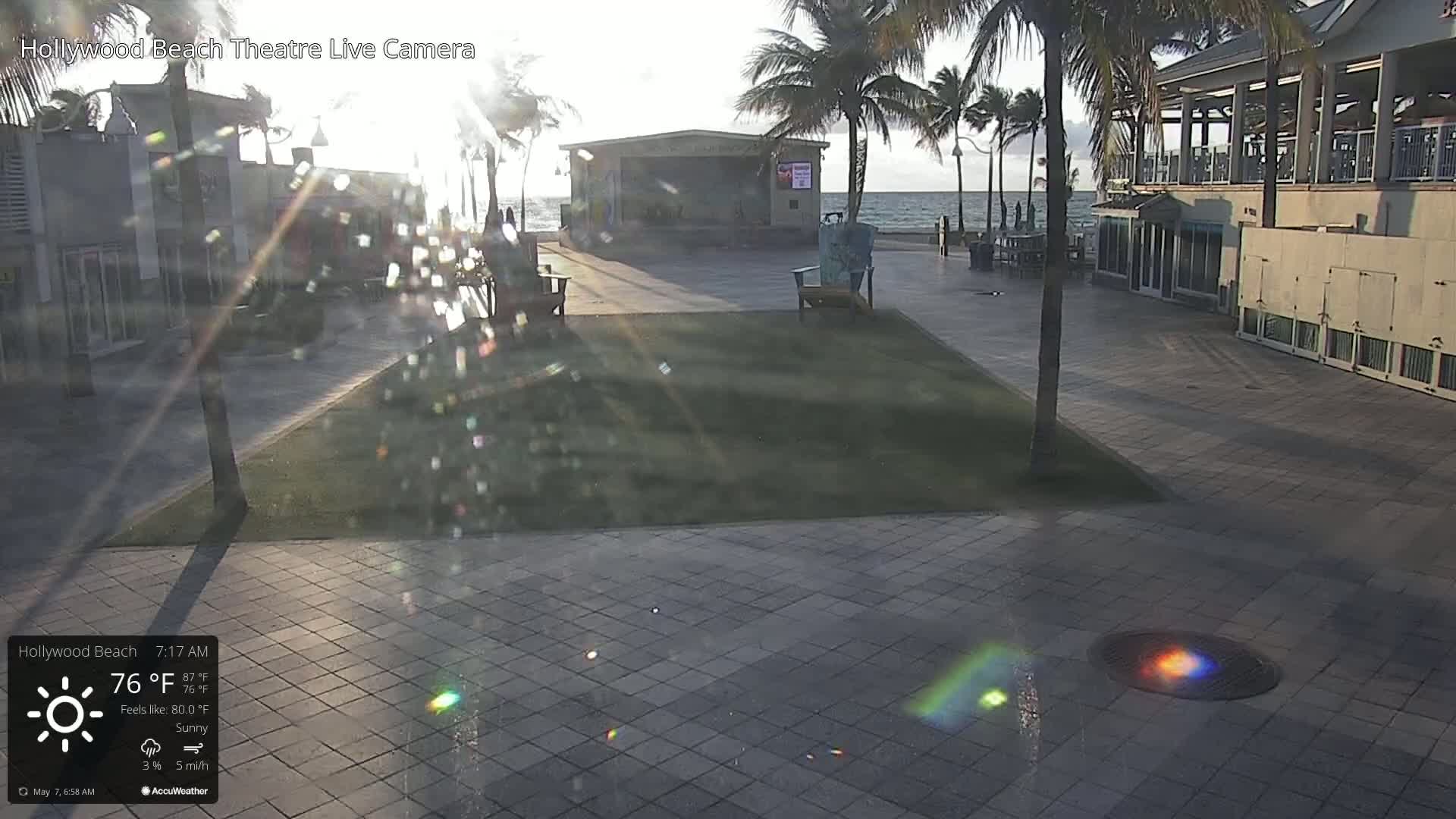 A sunny morning scene shows a paved plaza with palm trees, a small stage, and a building near a calm ocean.