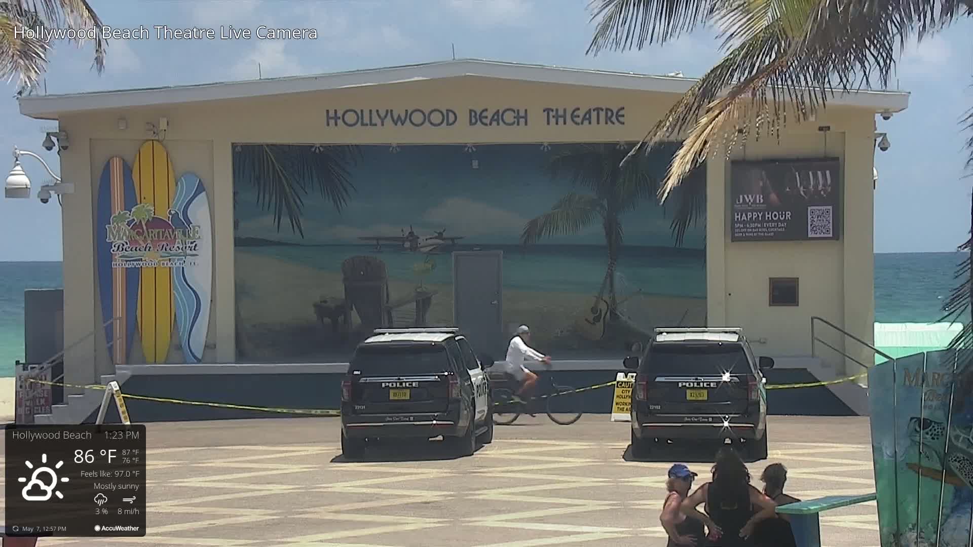 Under mostly sunny skies, two police SUVs are parked in front of a beachside theater with a mural depicting a tropical scene and surfboards; a person rides a bicycle nearby.