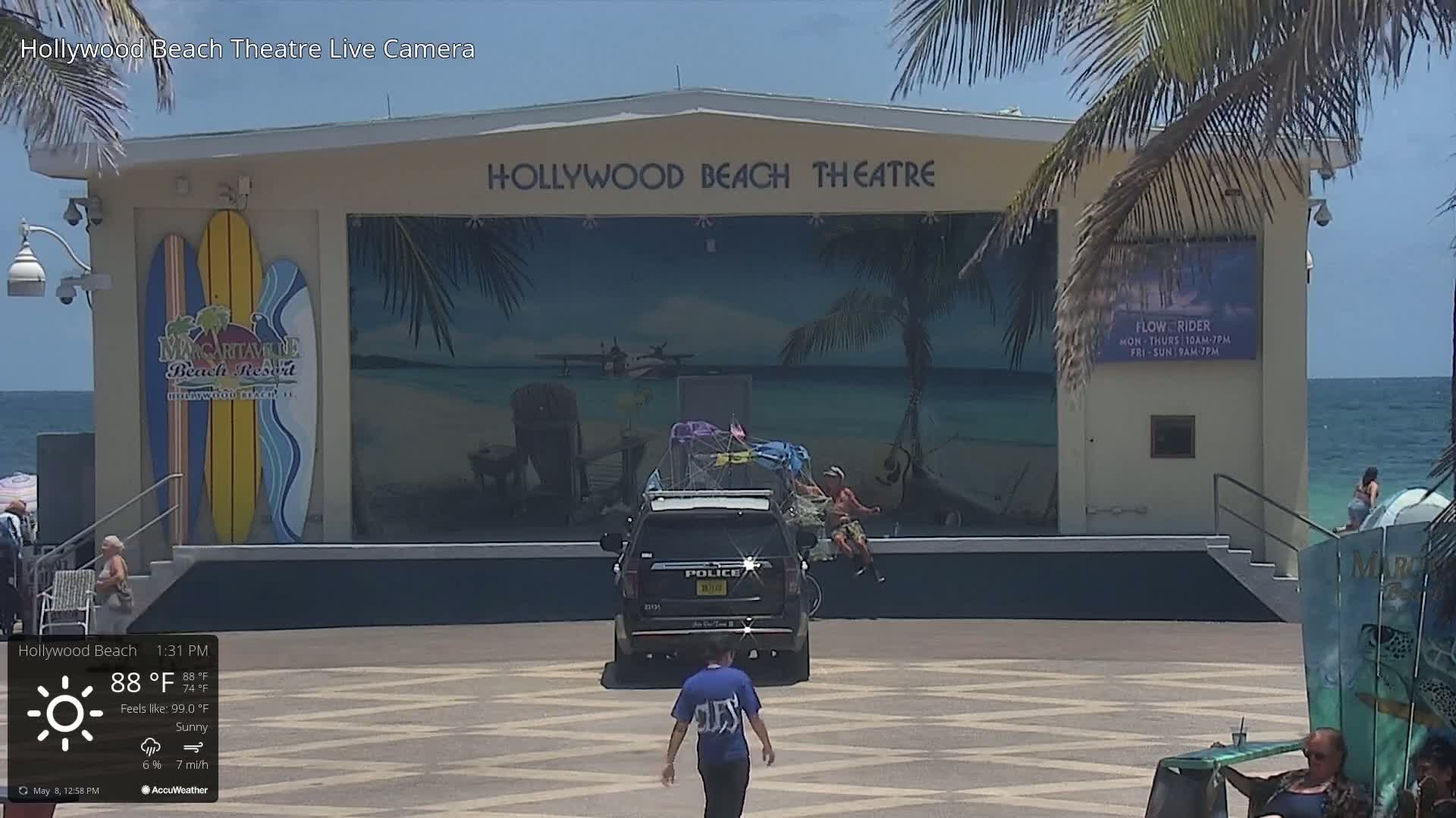 A sunny beach scene shows a beachside theatre with a mural depicting a tropical beach scene, a police SUV parked in front, and a few people walking around.