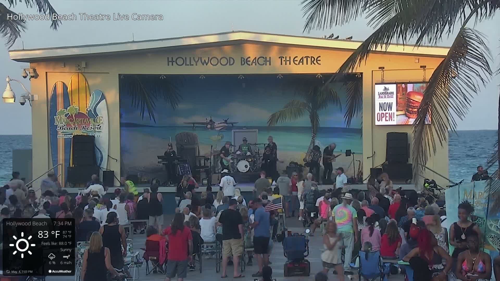 A large outdoor crowd watches a band perform on a stage at sunset under sunny skies.