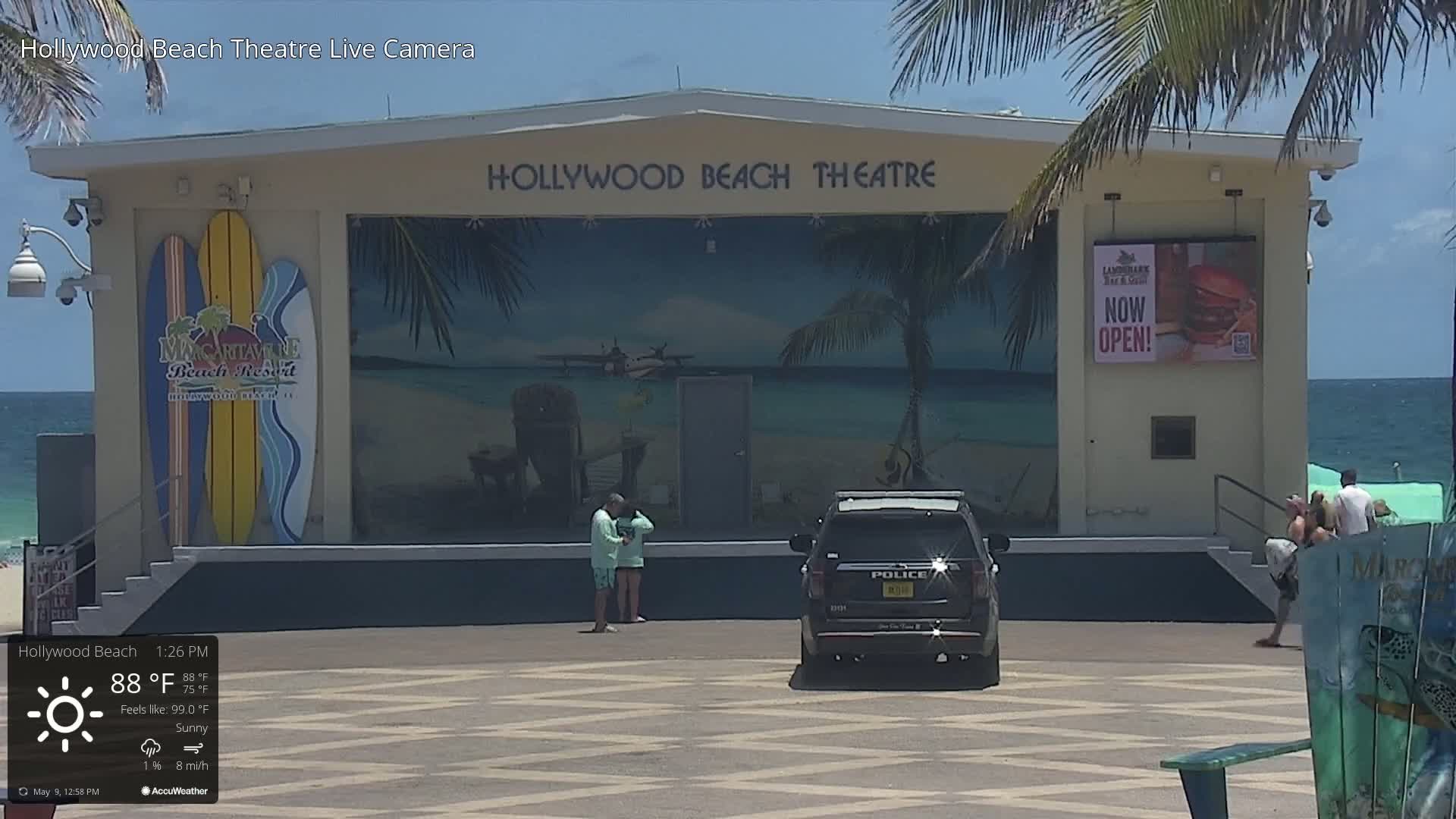 A sunny beach scene shows a police car parked in front of a beachside theater with a mural depicting a beach scene and surfboards; several people are near the theater.