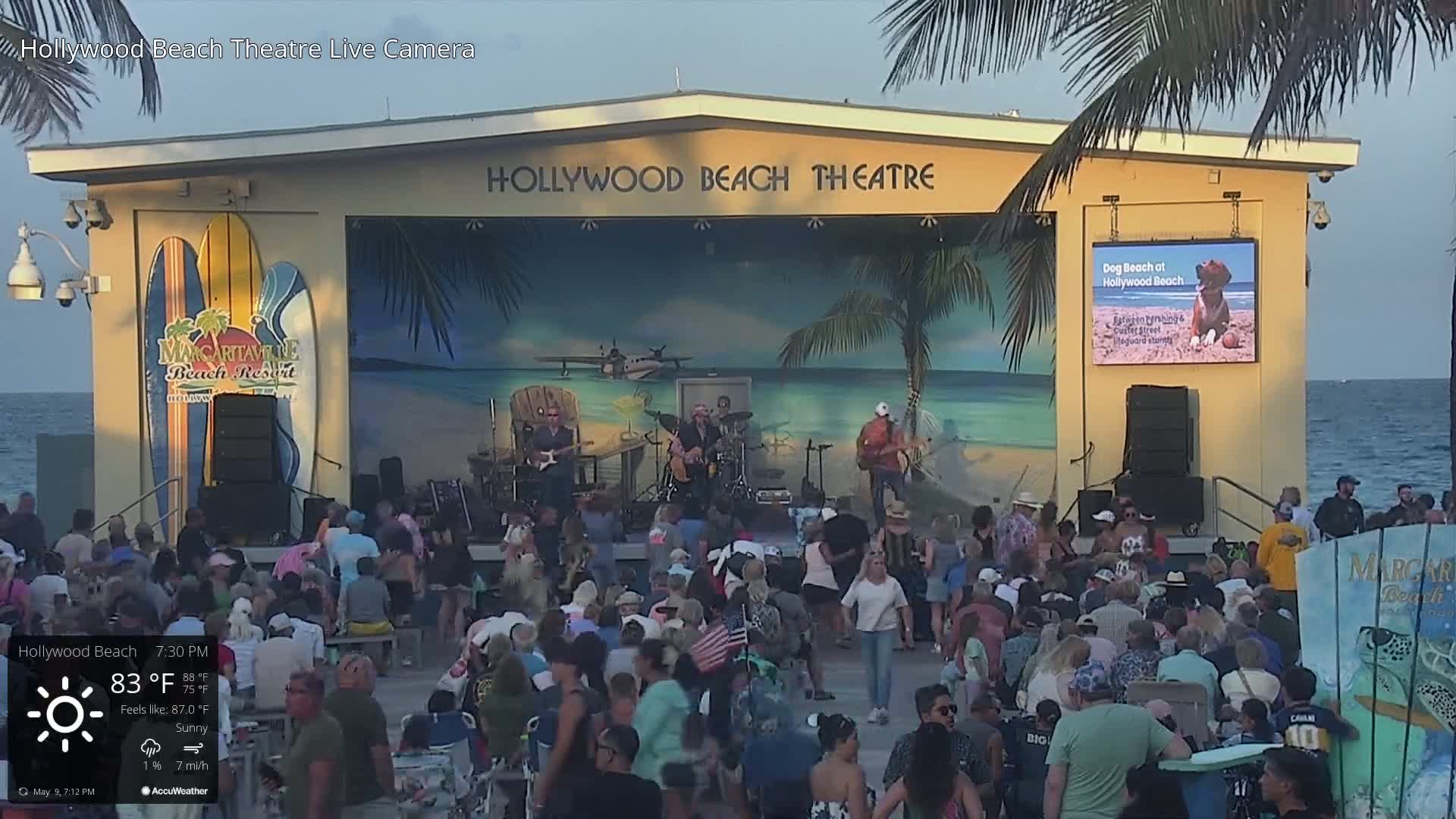 A large crowd watches a band perform on an outdoor stage on a sunny evening by the ocean.