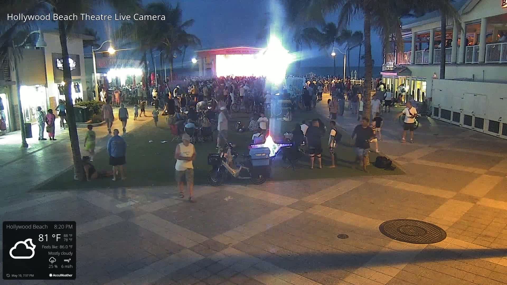 A nighttime outdoor scene shows a large crowd gathered around a stage on a paved area next to a beach, under mostly cloudy skies.