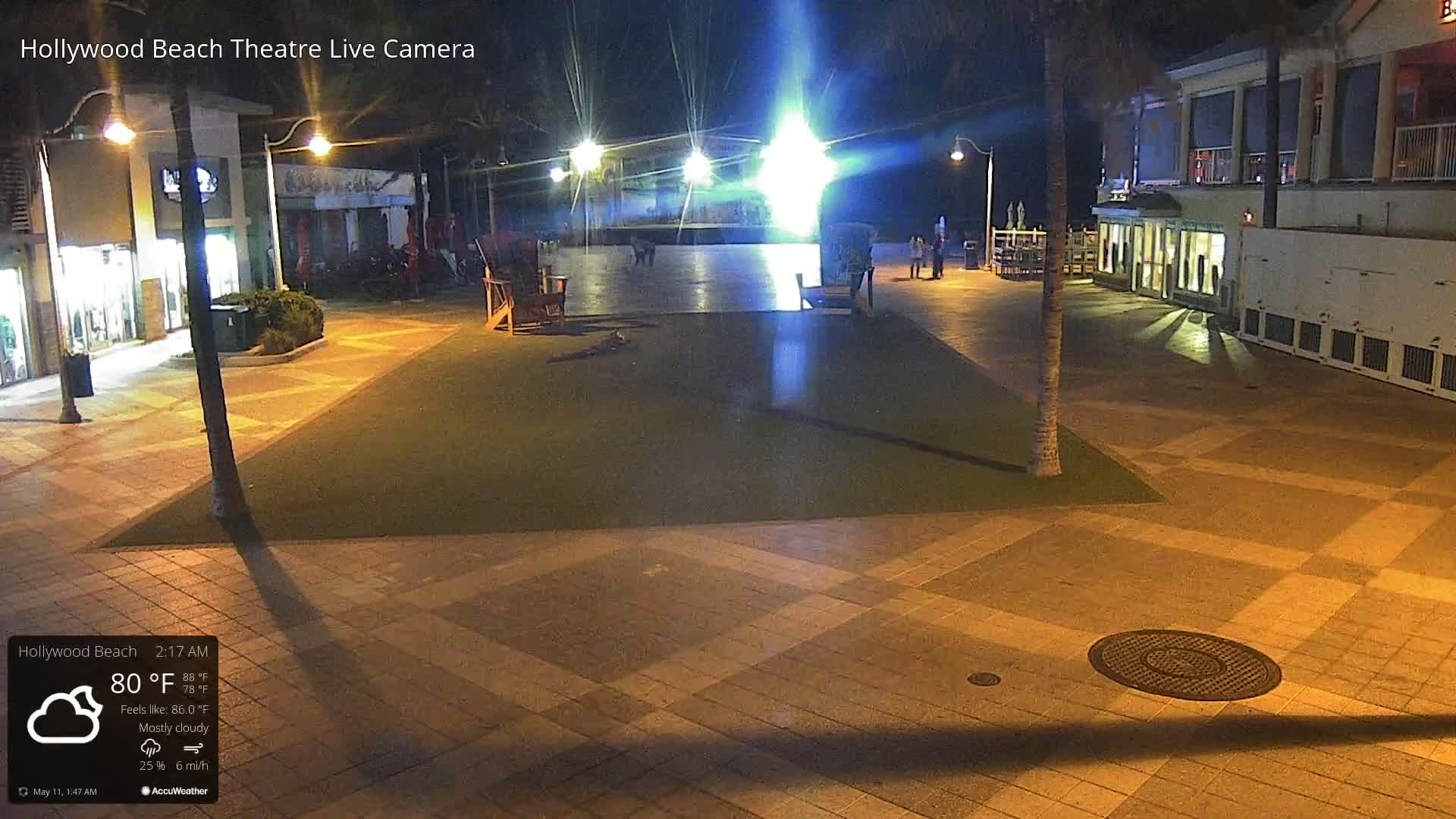 A mostly cloudy night scene shows a paved plaza with a few people and empty chairs under illuminated streetlights.