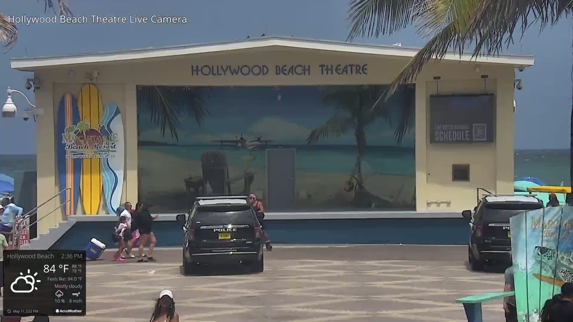 A mostly cloudy beach scene shows a beachside theater with people walking by, two police vehicles parked nearby, and a calm ocean in the background.