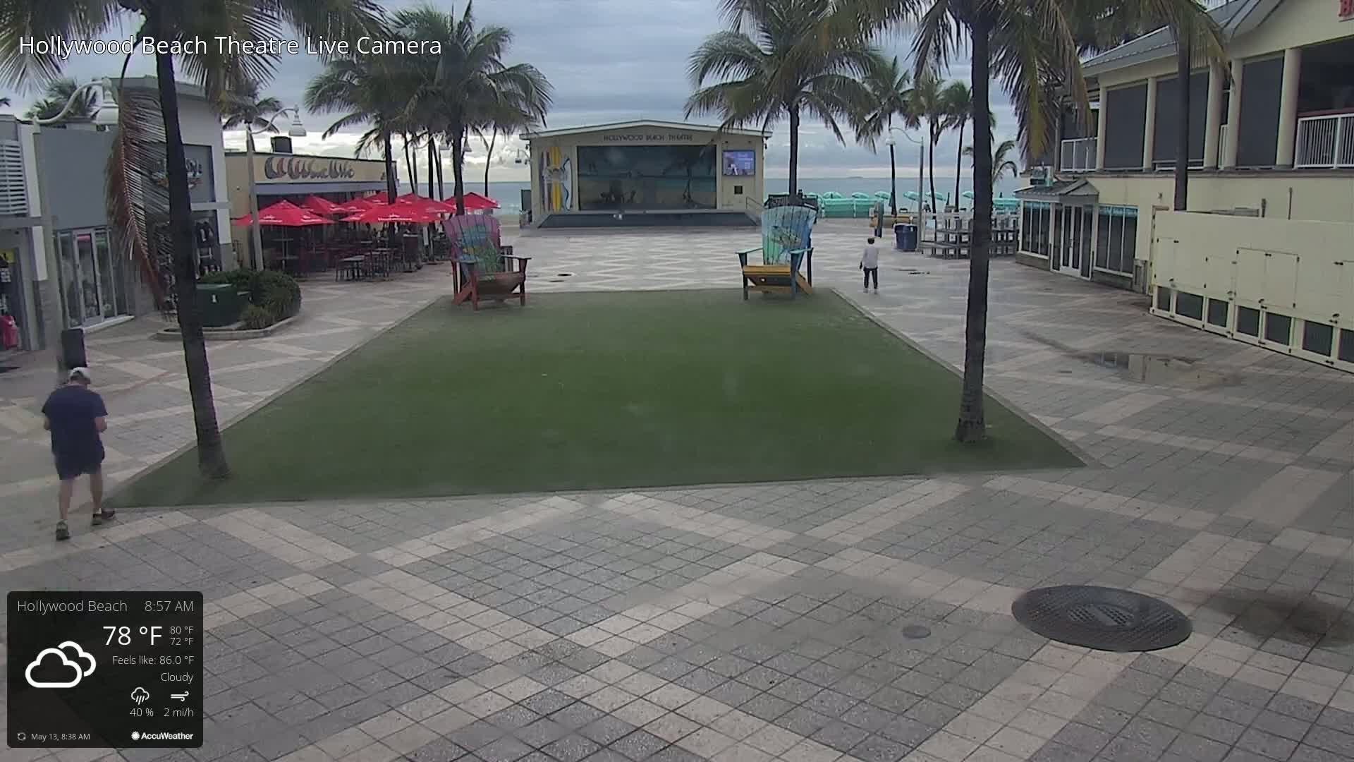 A cloudy morning scene shows a paved plaza with a grassy area, palm trees, a beachfront stage, and a few people walking.