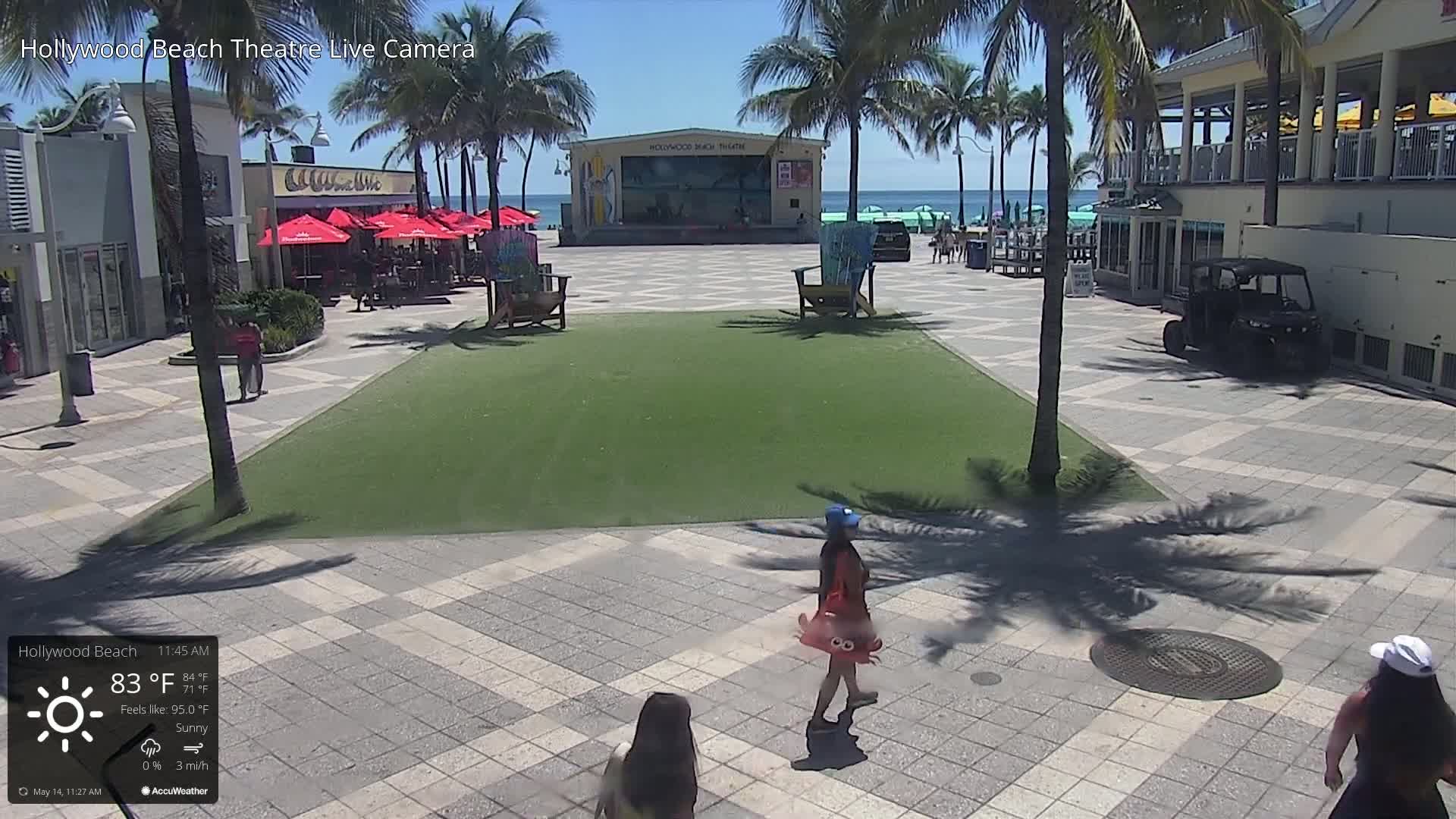 A sunny beachside plaza with a stage, palm trees, and people walking, under a bright, sunny sky.