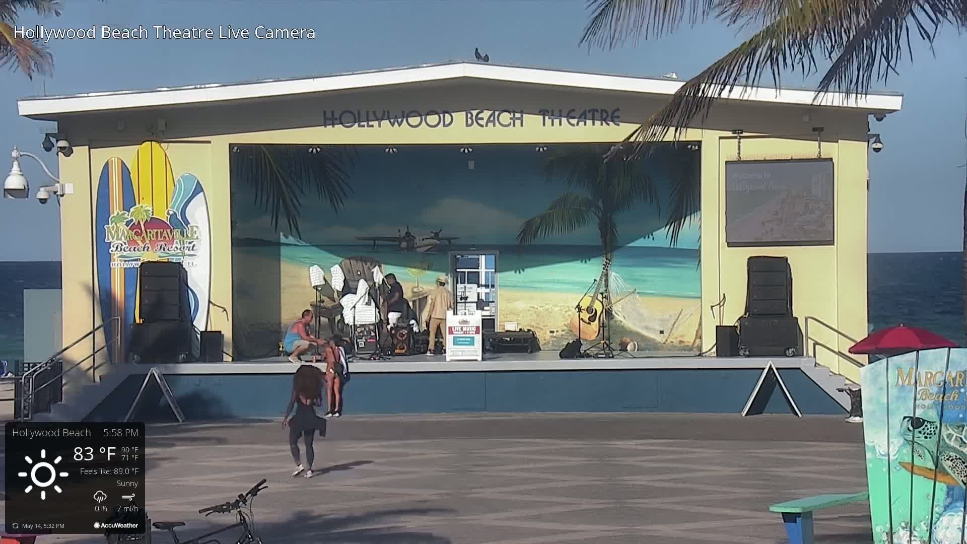 A beachside stage with a tropical mural and a small crowd of people under sunny skies.