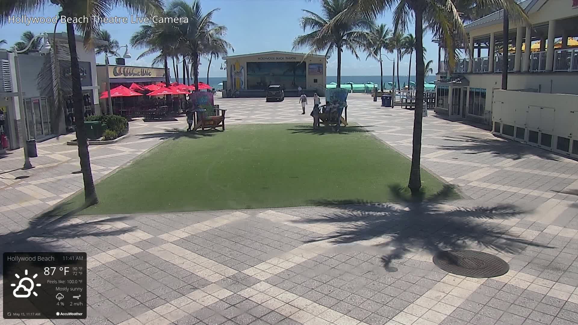 A mostly sunny beachside plaza with a grassy area, palm trees, and a stage is visible on a bright day.