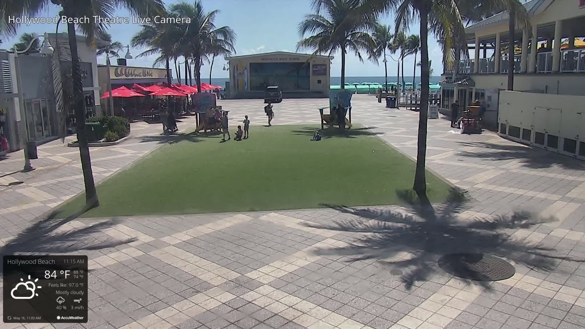 A mostly cloudy outdoor scene shows a paved plaza with a green grassy area in the center, several people, palm trees, beach umbrellas, buildings, and a stage near the ocean.