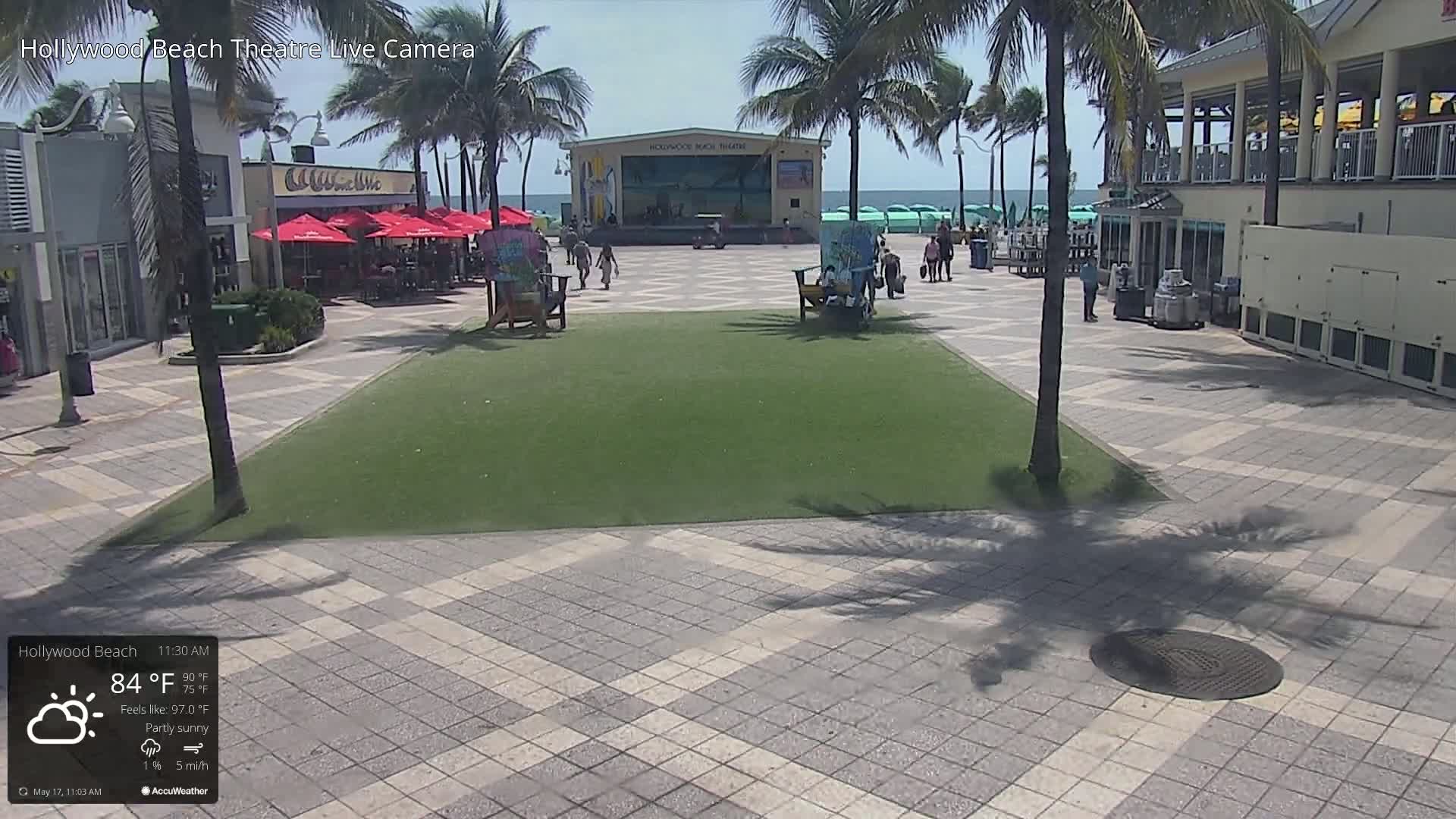 A paved plaza with a grassy area in the center, featuring an outdoor stage near an ocean, under partly sunny skies.