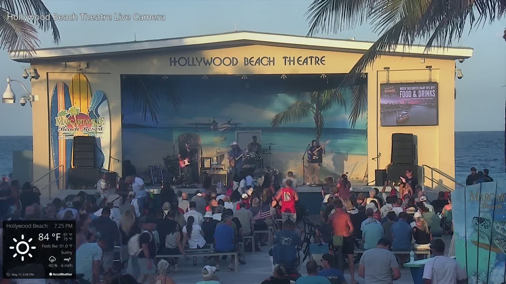 A band plays on a beachside stage in front of a seated audience under sunny skies.