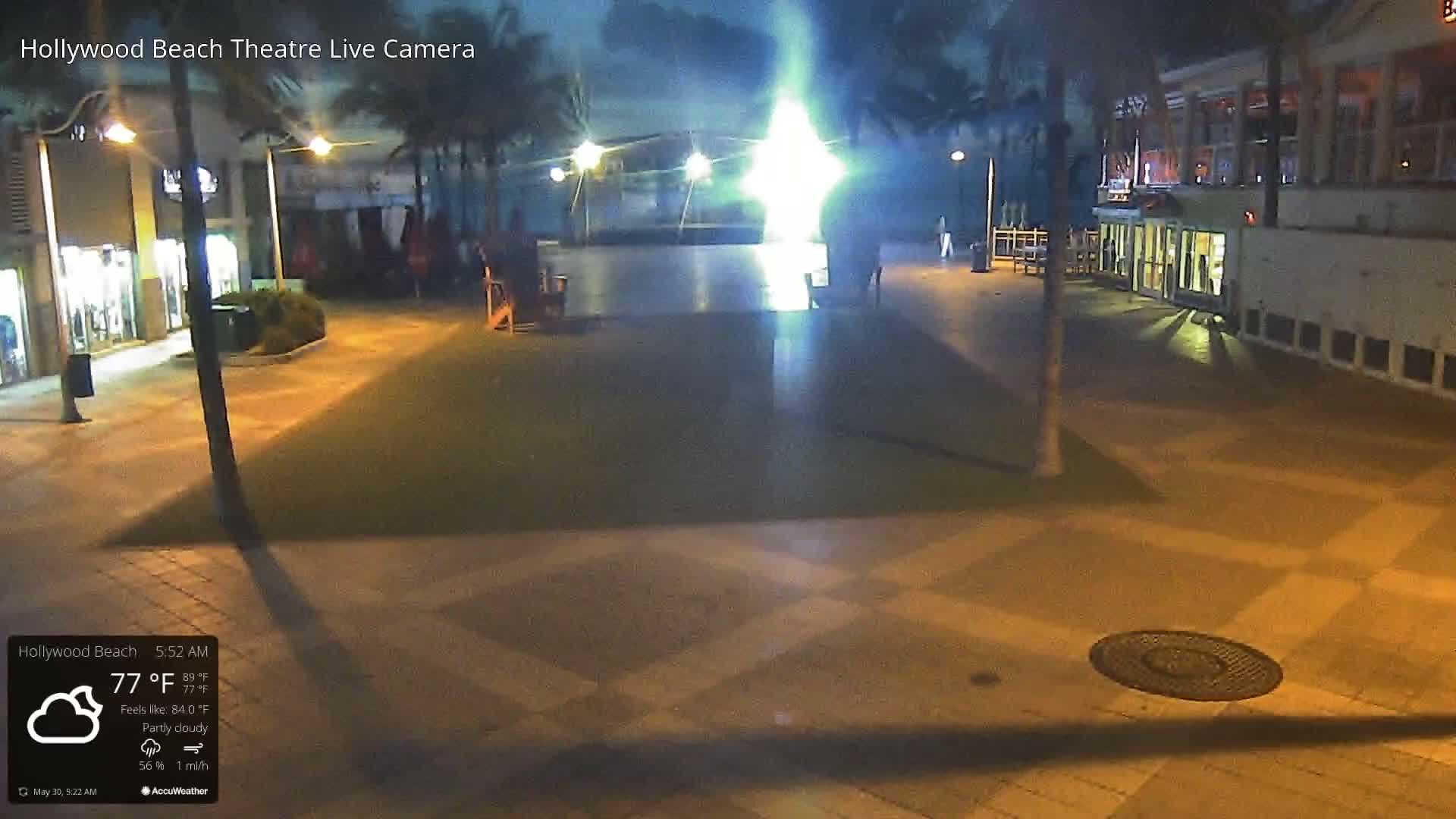 A nighttime, partly cloudy scene shows an empty plaza with buildings and palm trees, illuminated by streetlights.