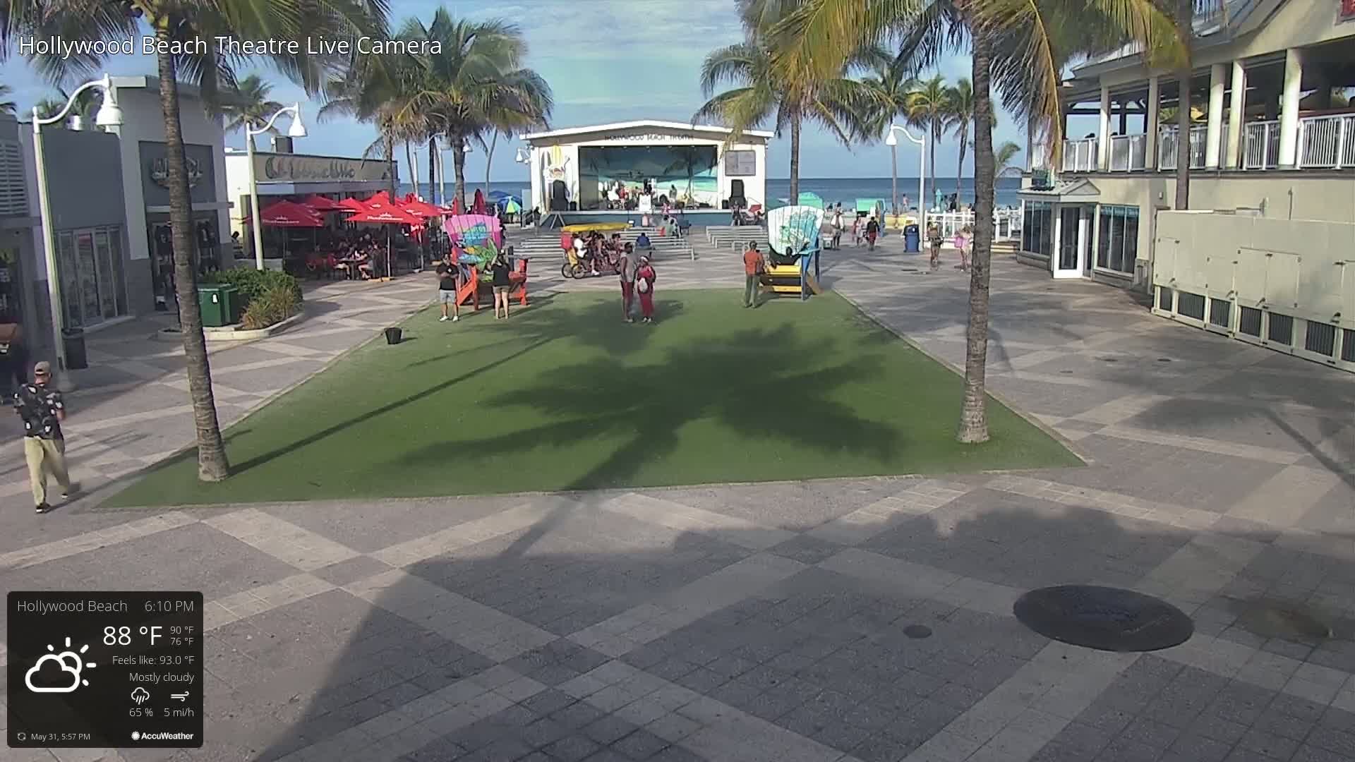 A mostly cloudy beachside plaza with a stage, palm trees, and people, under a mostly cloudy sky with a temperature of 88°F.