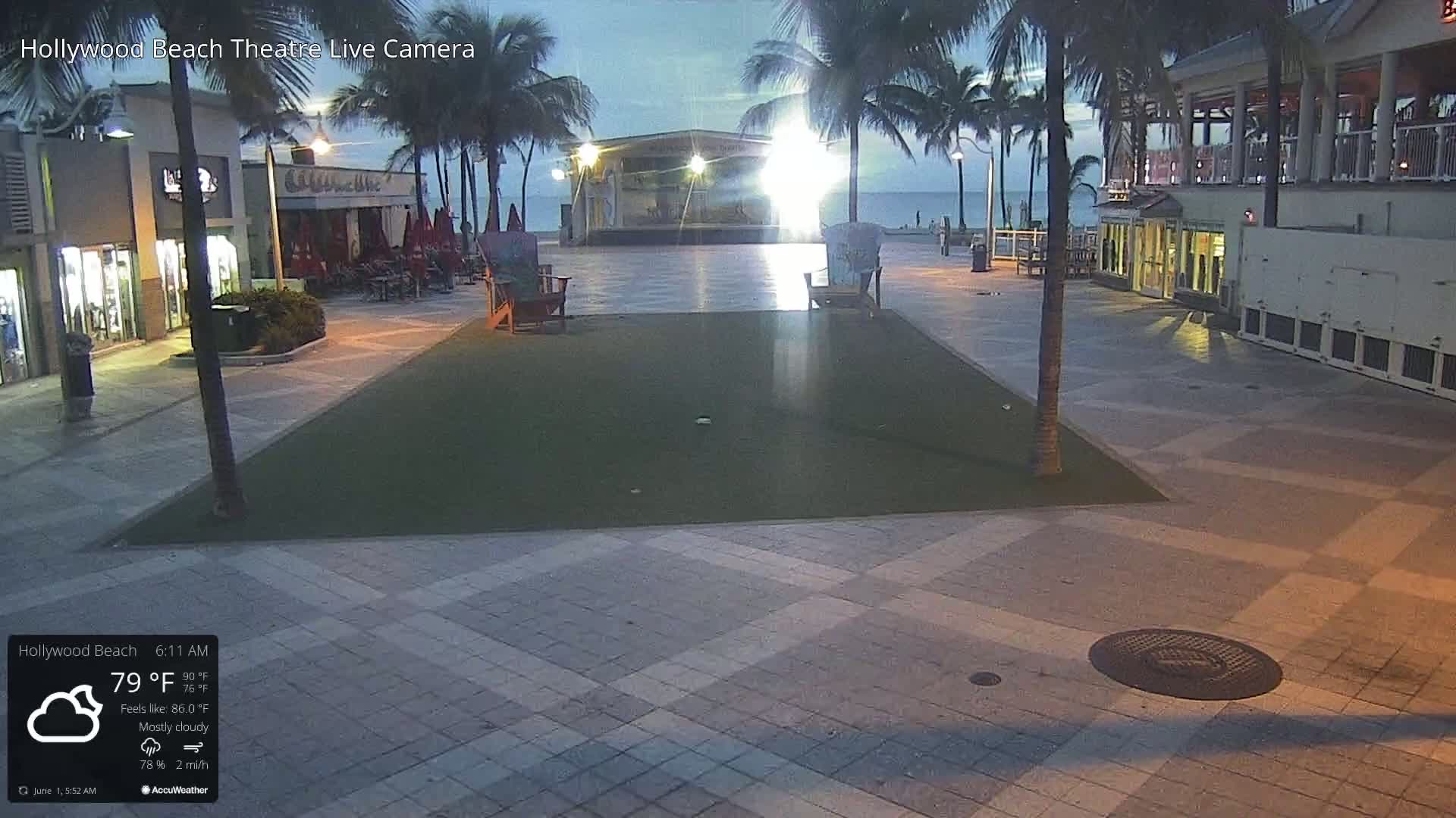 An empty plaza with a grassy area, stage, and beach visible in the background under mostly cloudy skies at night.