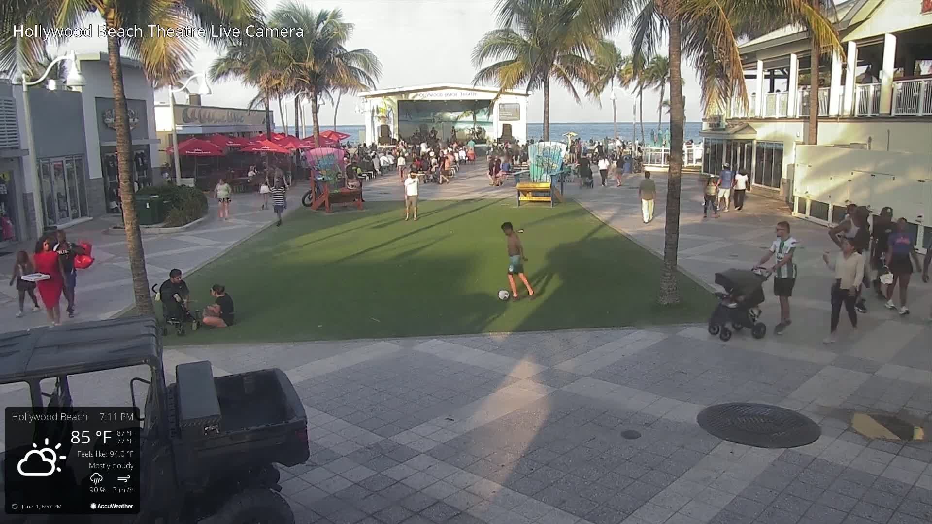 A mostly cloudy outdoor scene shows a paved plaza with a grassy area, people walking, and a boy kicking a soccer ball near a beachfront stage with a small crowd.