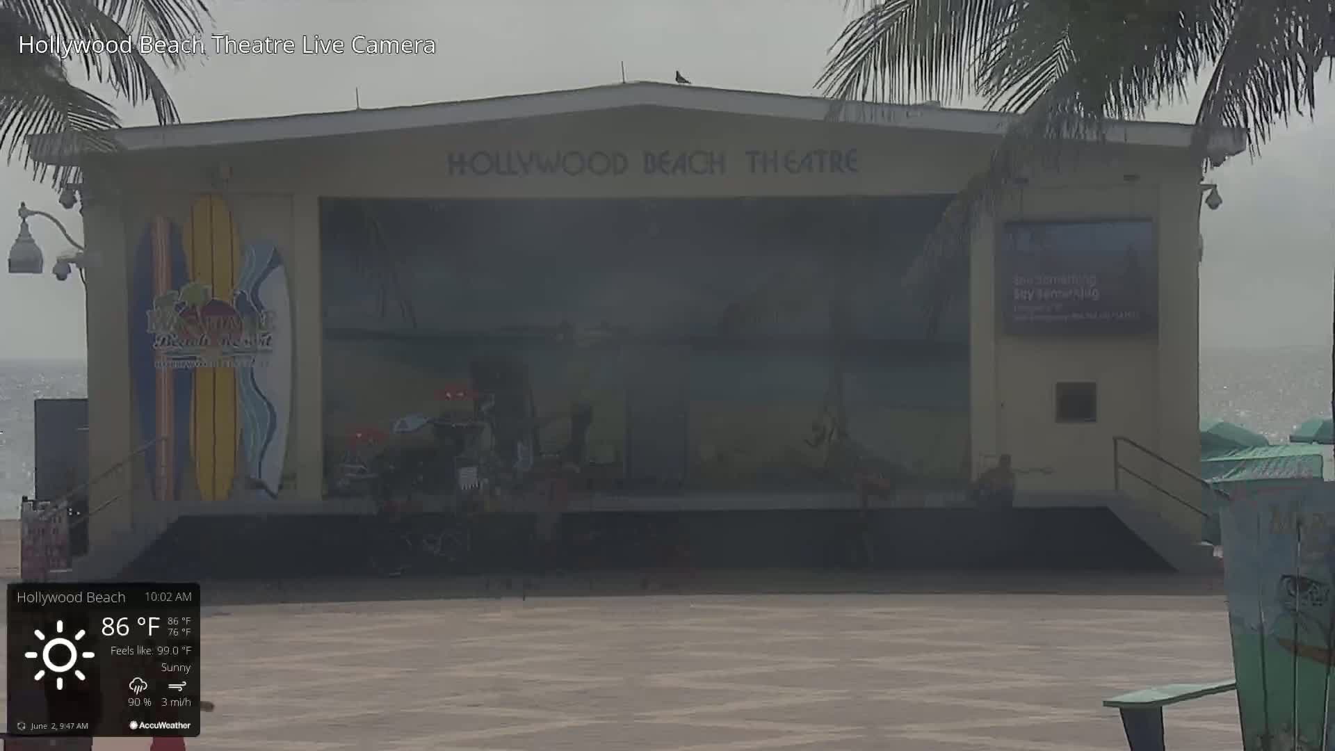 A hazy, sunny outdoor view of a beachside theatre with a stage featuring a mural of surfboards.