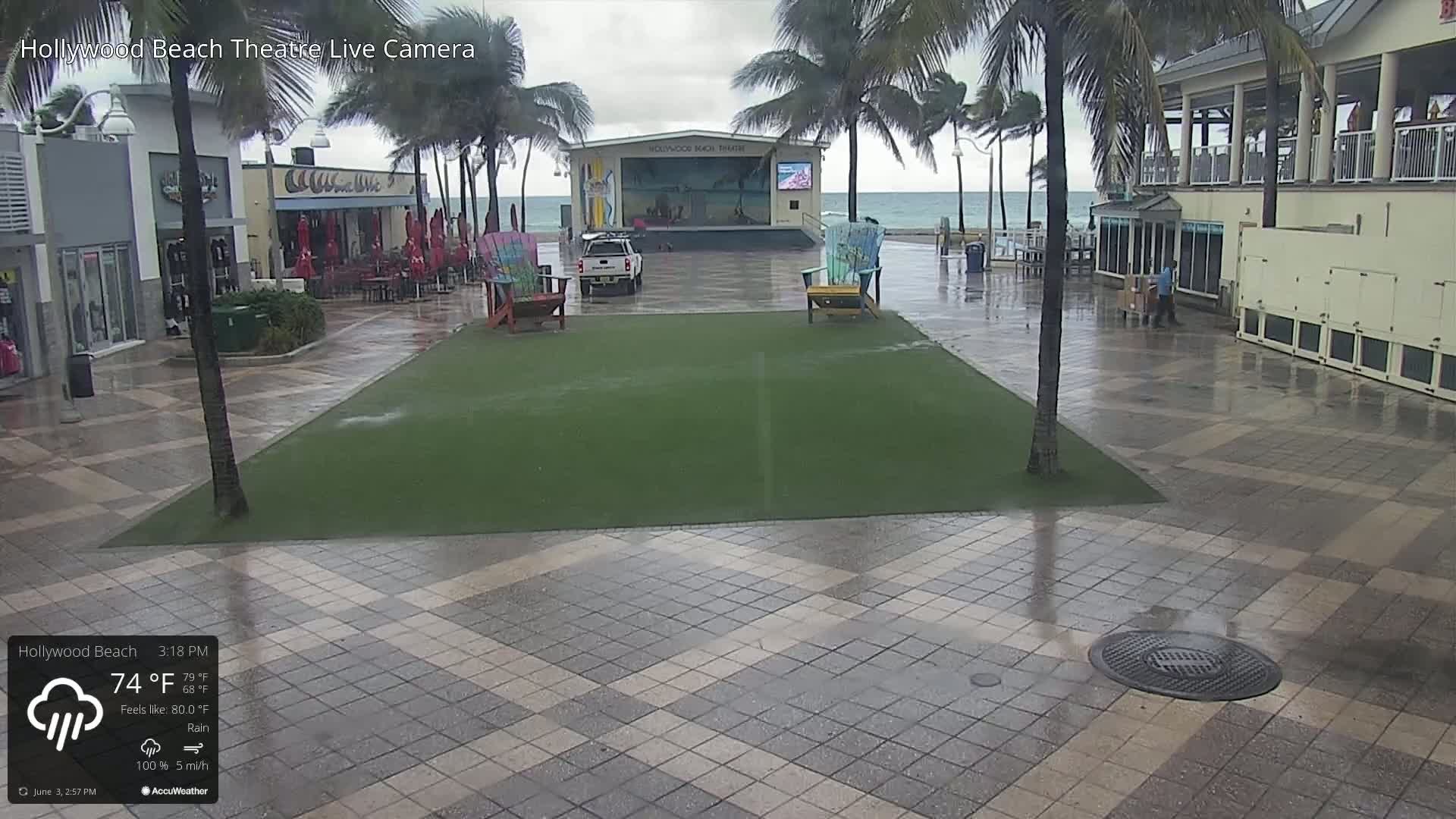 A wet paved plaza with a grassy area in the center, an outdoor stage behind it, palm trees, and ocean in the background under rainy conditions.