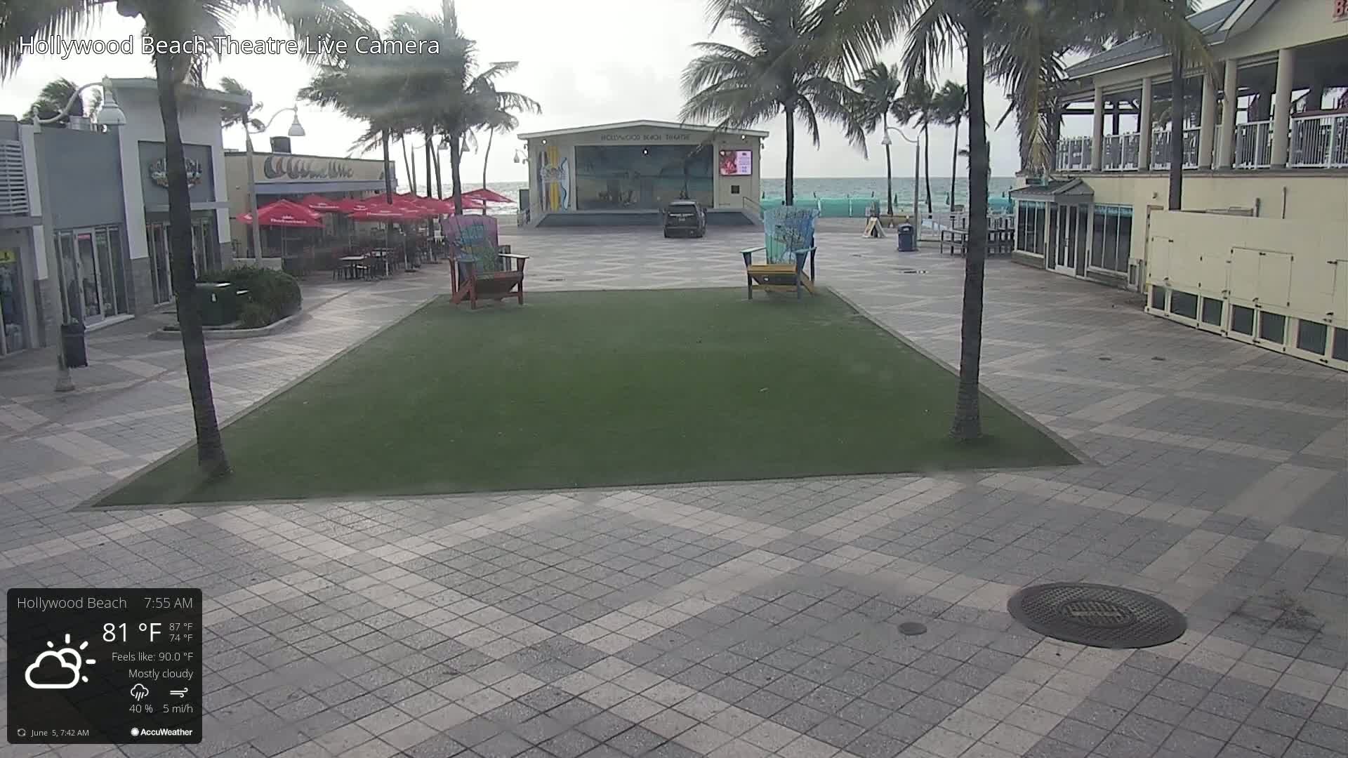 A mostly cloudy outdoor scene shows a paved plaza with a patch of artificial turf, palm trees, beach-side buildings, and an ocean in the background.