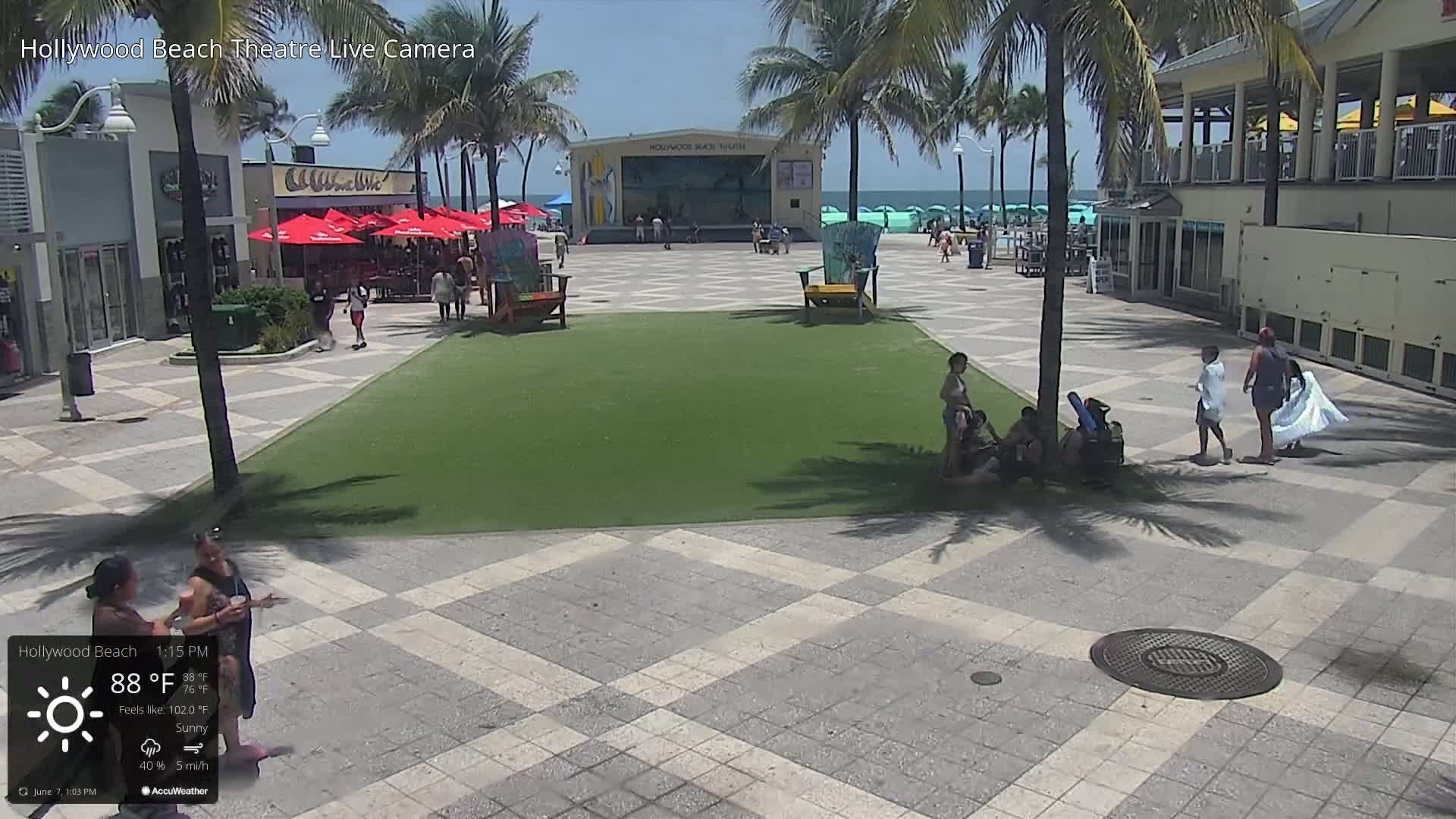A sunny beachfront plaza with a stage, palm trees, and several people scattered about.