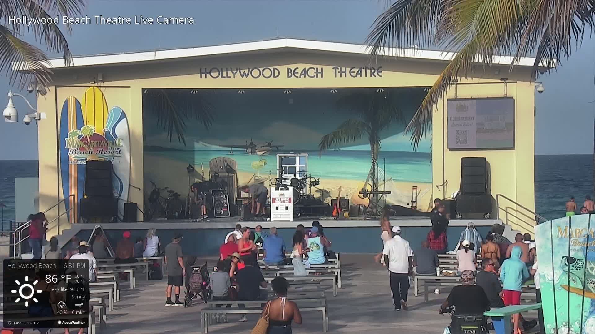 A sunny outdoor beachside theater with a band playing on stage and an audience seated at picnic tables.