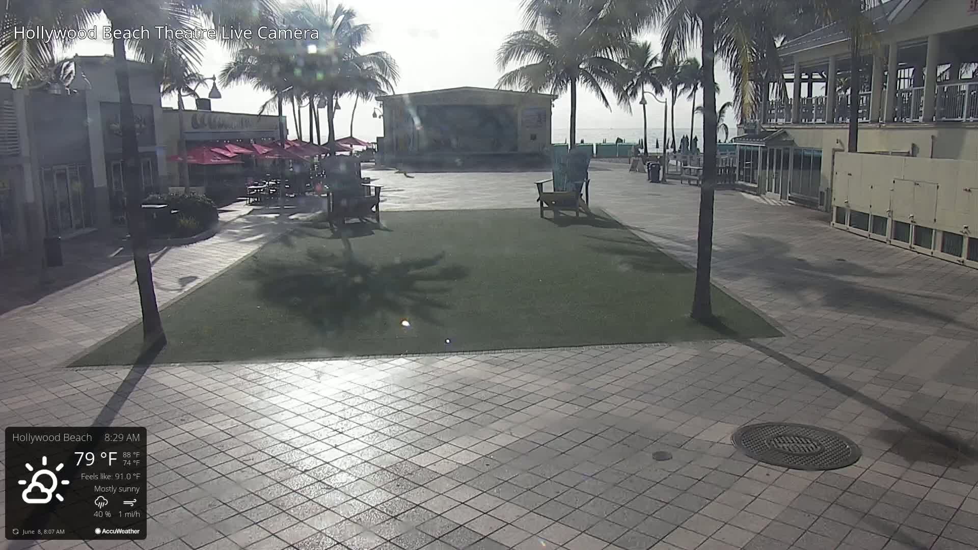 A mostly sunny outdoor scene shows a paved plaza with a stage, palm trees, and buildings, under a bright sky.