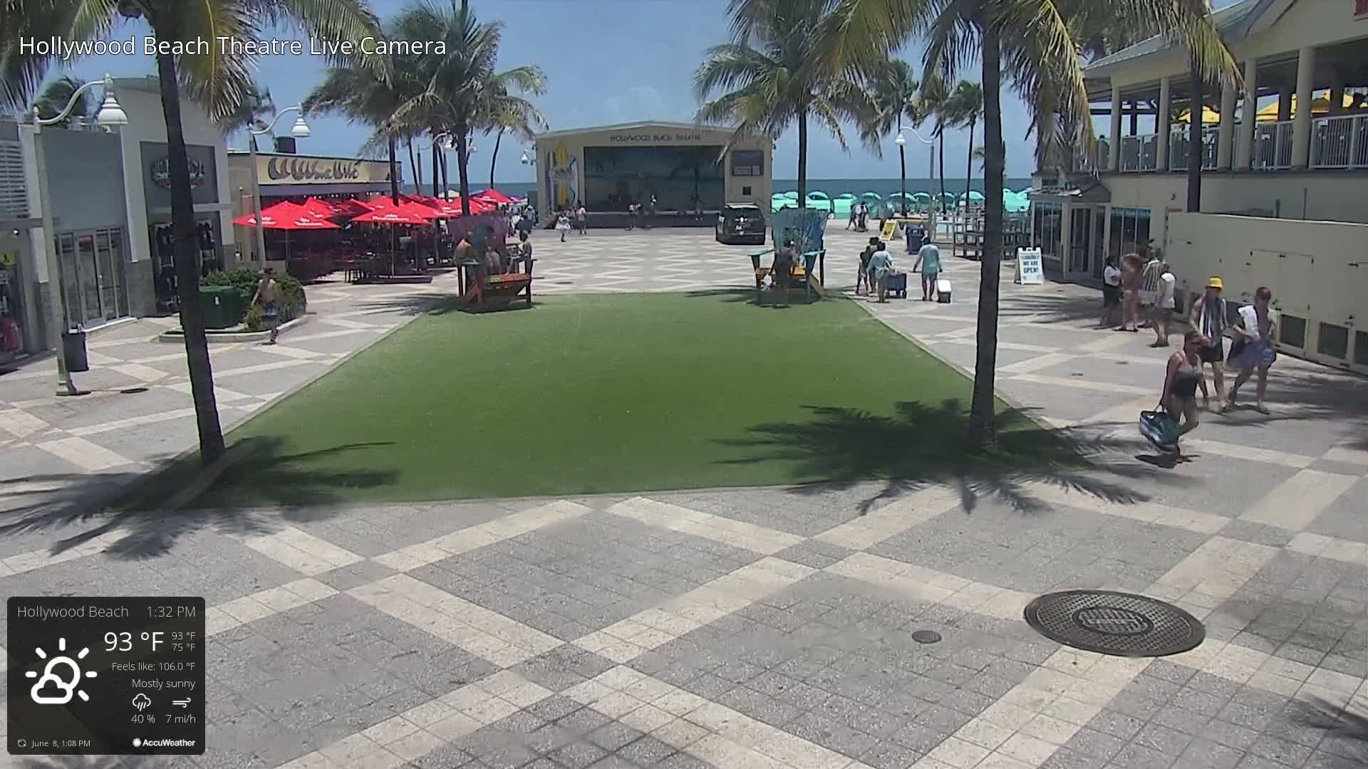 A mostly sunny beachside plaza with a stage, palm trees, and several people walking around on a bright, hot day.