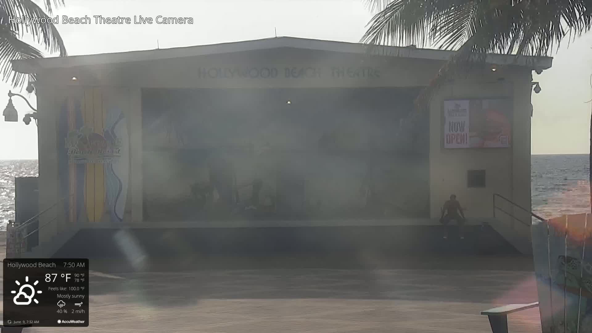 A mostly sunny outdoor scene shows the Hollywood Beach Theatre, with a person sitting on the ground in front of it, next to a calm ocean.