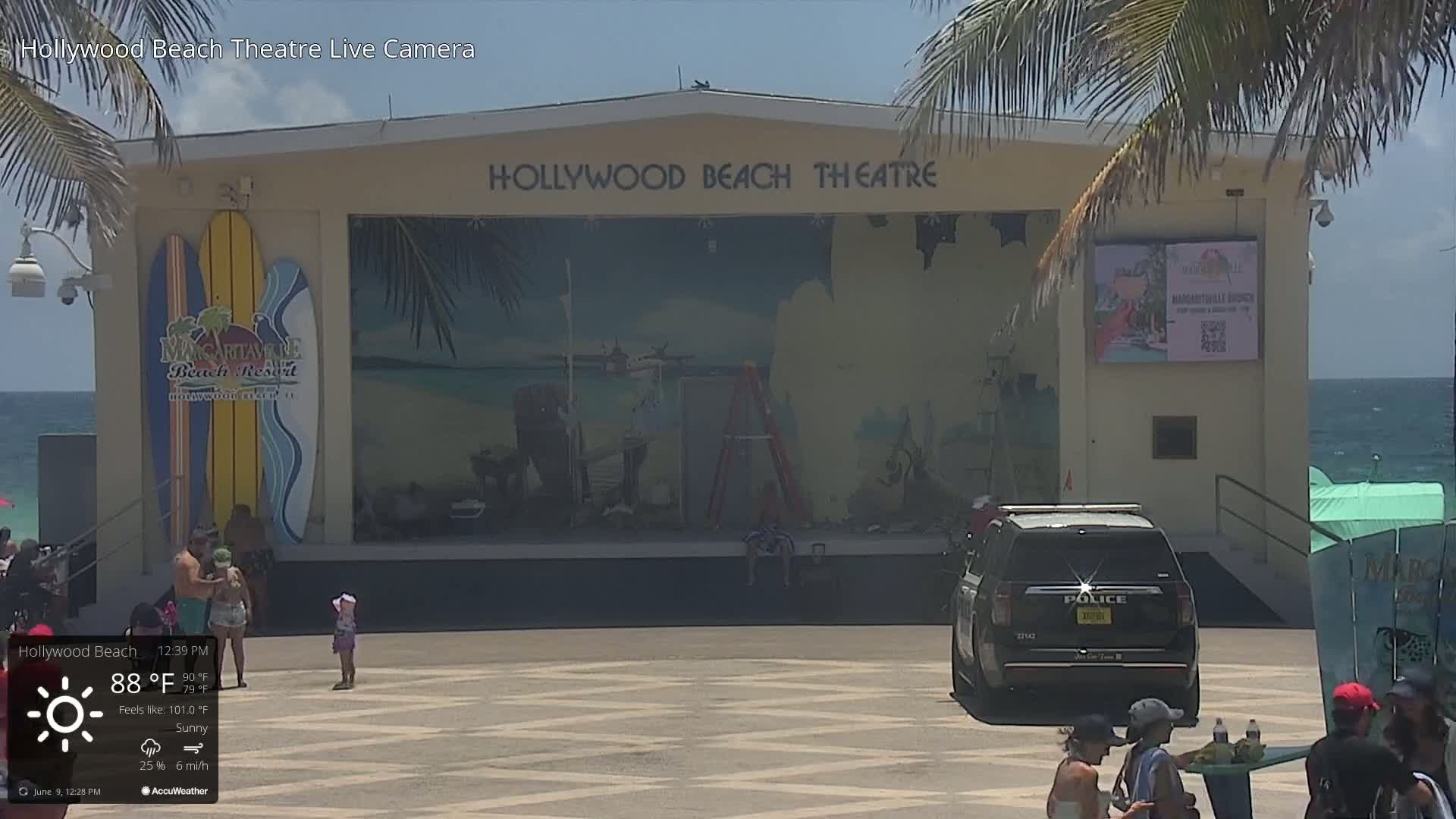 A sunny, outdoor scene shows a beachside theater with a mural, a police car parked in front, and several people on the plaza.