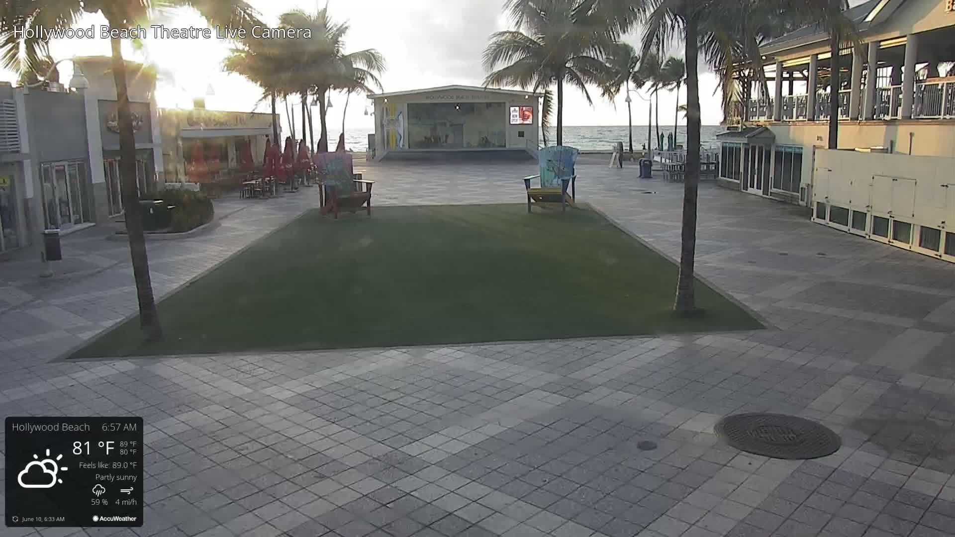 A partly sunny morning scene shows a paved plaza with a small patch of grass, two Adirondack chairs, a beachfront stage, and palm trees under a mostly clear sky.