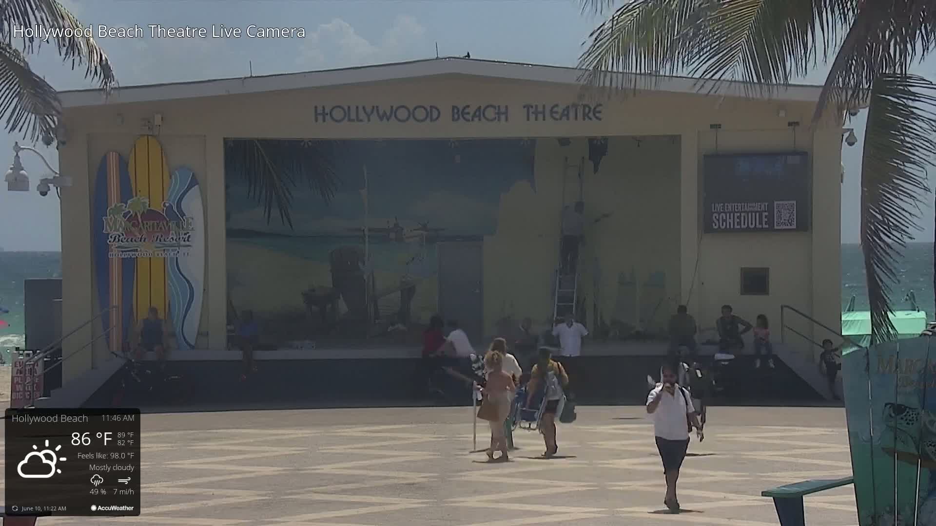 A mostly cloudy day with a temperature of 86°F shows people walking in front of an outdoor stage with beach-themed murals and signage.