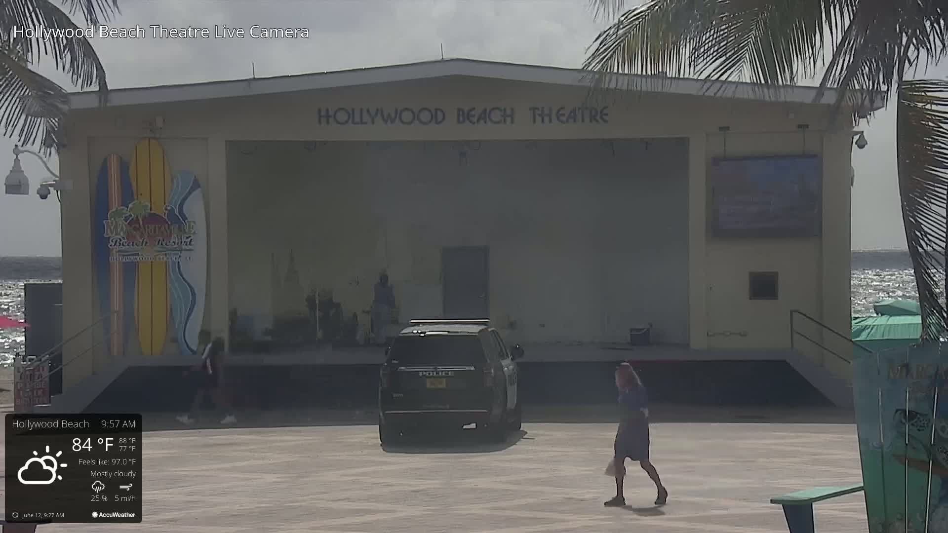 A mostly cloudy outdoor scene shows a mostly empty stage at the Hollywood Beach Theatre, with a police vehicle parked nearby, and a few people walking on the boardwalk.