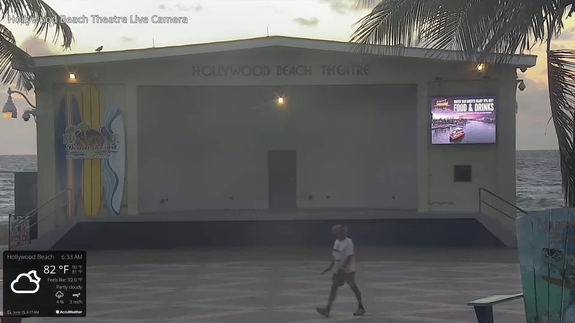 A mostly empty outdoor stage area on a partly cloudy morning, with a man walking on the concrete, next to the ocean.