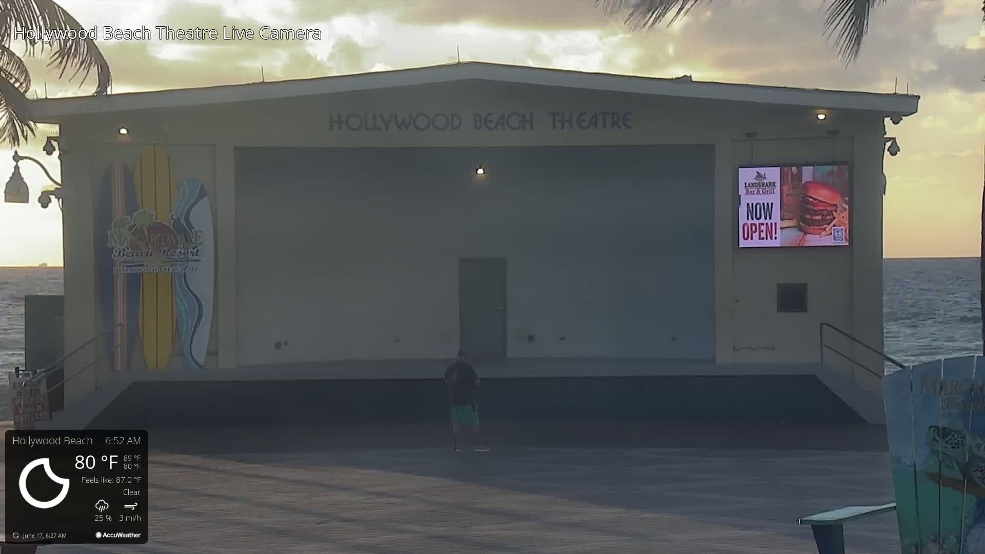 An outdoor beachfront stage, under a clear sunrise sky, with a single person standing on the stage.