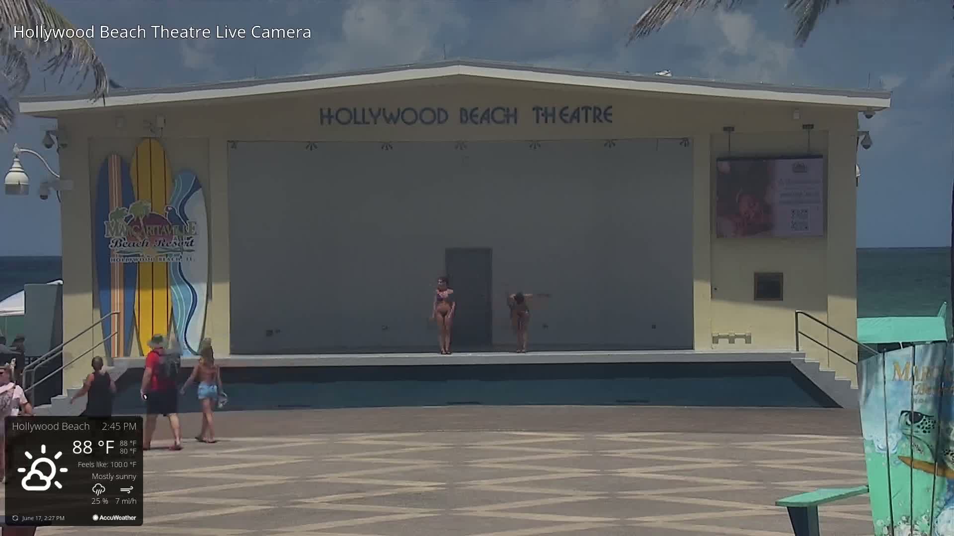 An outdoor mostly sunny scene shows two women in bikinis standing on a stage in front of the Hollywood Beach Theatre, with several people walking around the area near the ocean.