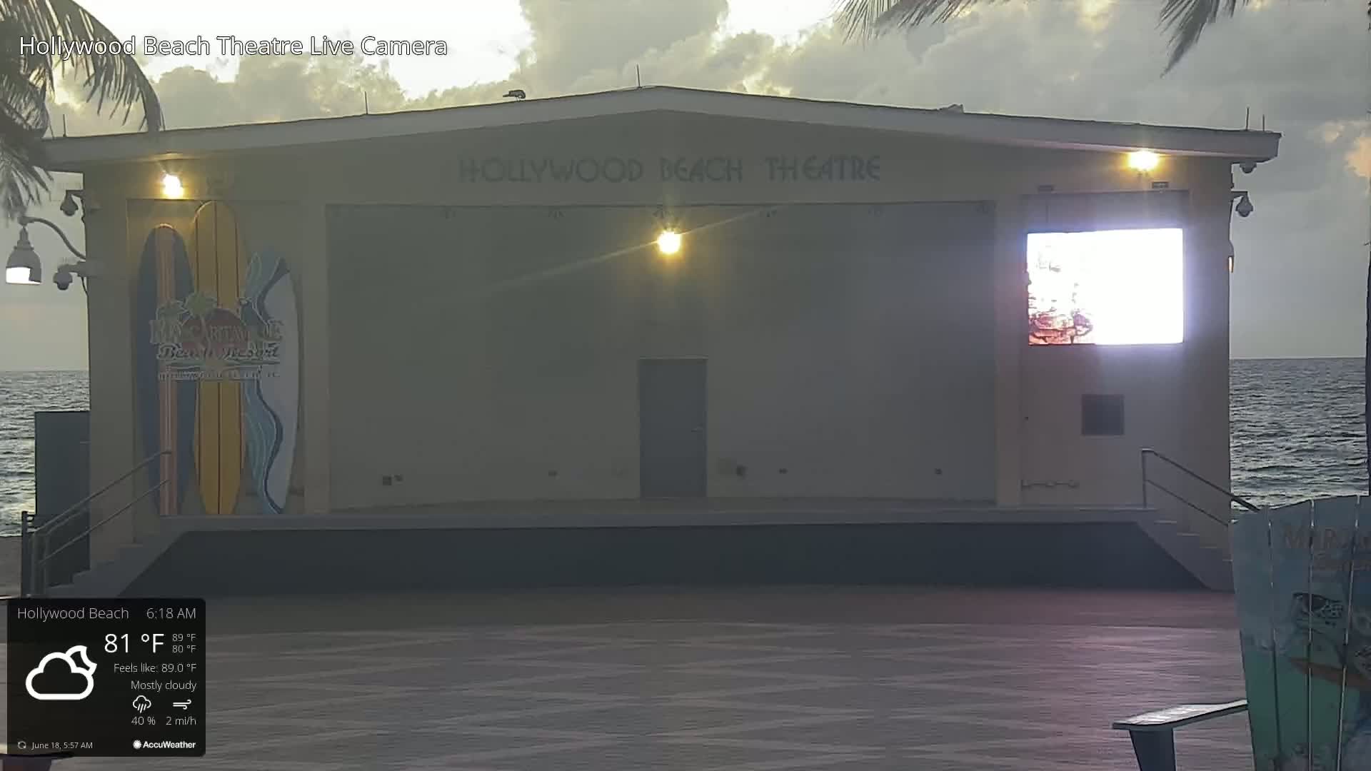 A mostly cloudy sunrise illuminates a beachside theatre with a large screen displaying a bright image; the ocean is visible in the background.