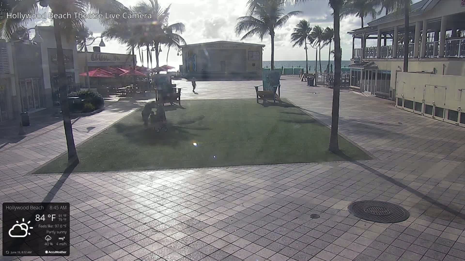 A partly sunny outdoor scene shows a paved plaza with a grassy area, palm trees, beach-side buildings, and a few people.