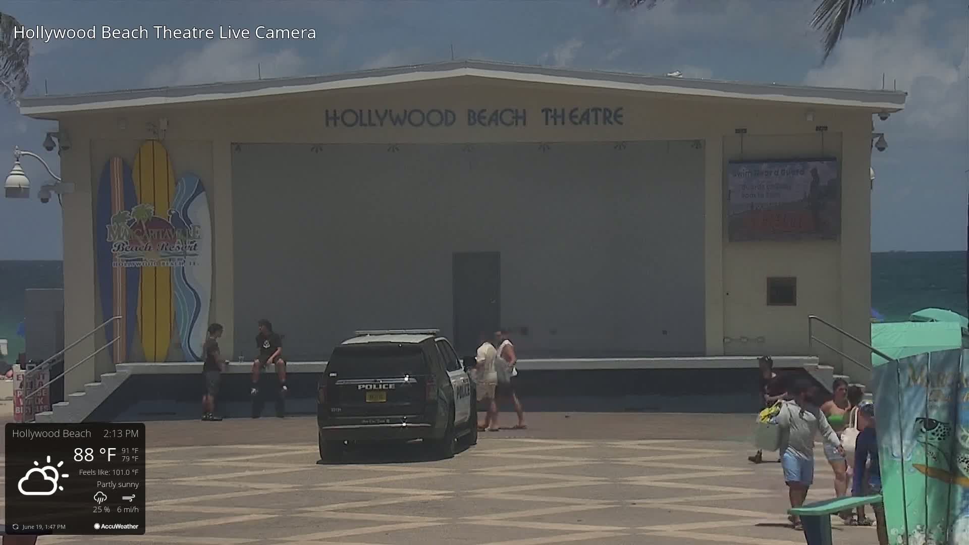A partly sunny day at the Hollywood Beach Theatre, with a police vehicle parked in front of the stage where several people are standing or walking.