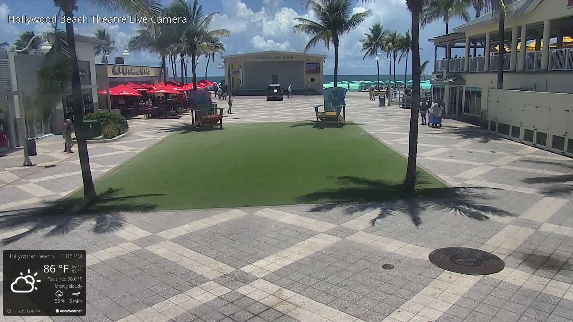 A mostly cloudy outdoor scene shows a paved plaza with a grassy area, beach chairs, palm trees, a beachfront theater, and a restaurant, with a few people visible.