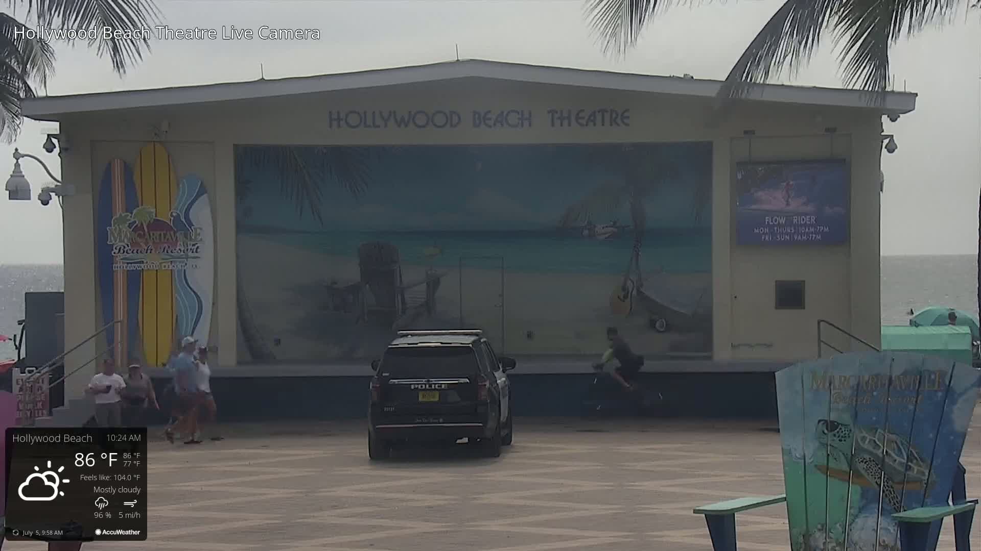 A mostly cloudy day at 86°F shows a police SUV parked in front of the Hollywood Beach Theatre, where several people are walking and a person is riding a bicycle.