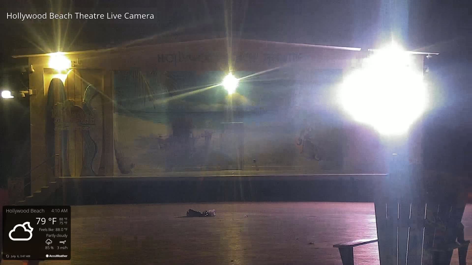 At night, under partly cloudy skies, an empty stage with painted murals is brightly lit, and a single chair sits in the foreground.