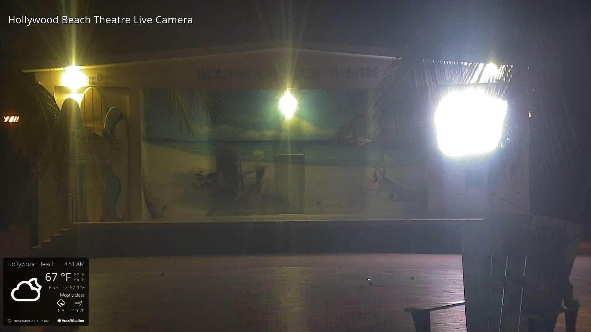 On a clear night, an outdoor stage featuring a beach-themed mural is brightly lit, with an Adirondack chair positioned on a dark paved surface in the foreground.