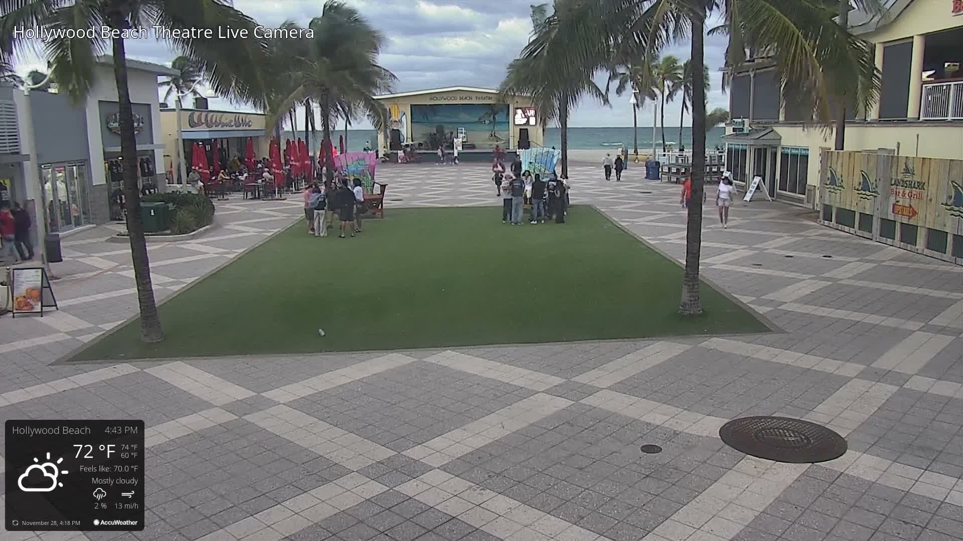 A bustling beachside promenade is visible with numerous people walking and gathering under palm trees, alongside shops and a stage, with the ocean in the background, all under a mostly cloudy sky at 72°F.