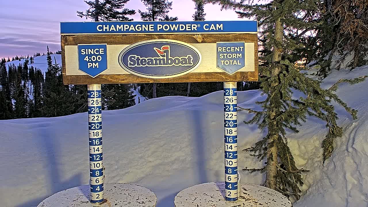 A snow depth gauge and sign indicating recent snowfall are situated in a snowy landscape at dusk, with evergreen trees in the background.