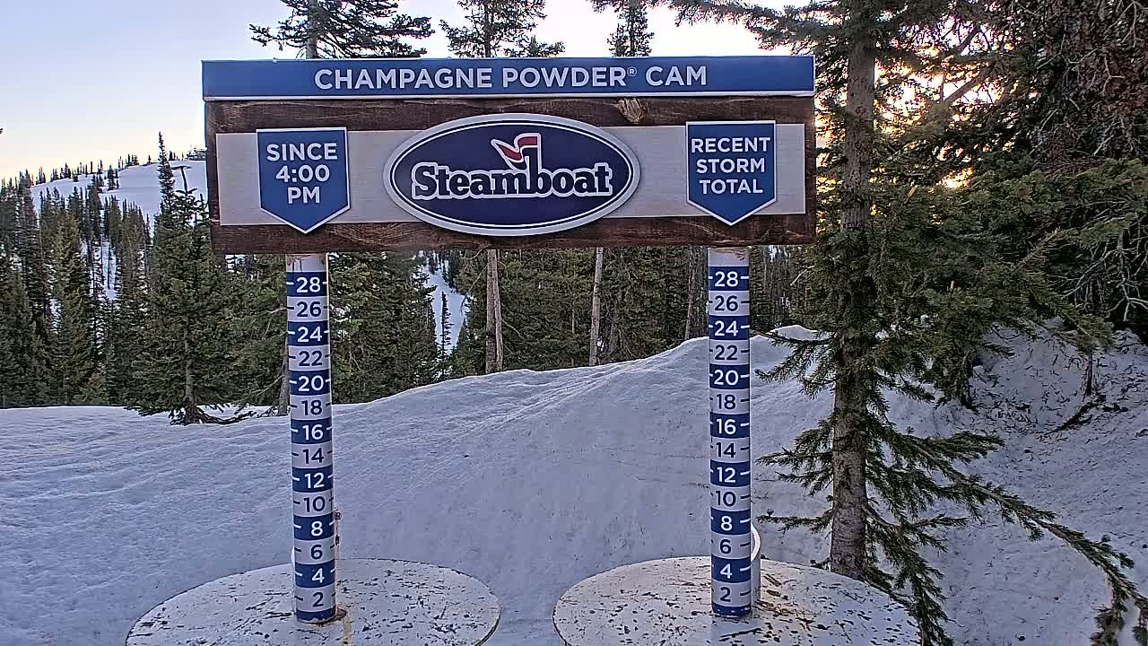 A snow depth gauge and sign stand in a snowy landscape at sunset, surrounded by evergreen trees.